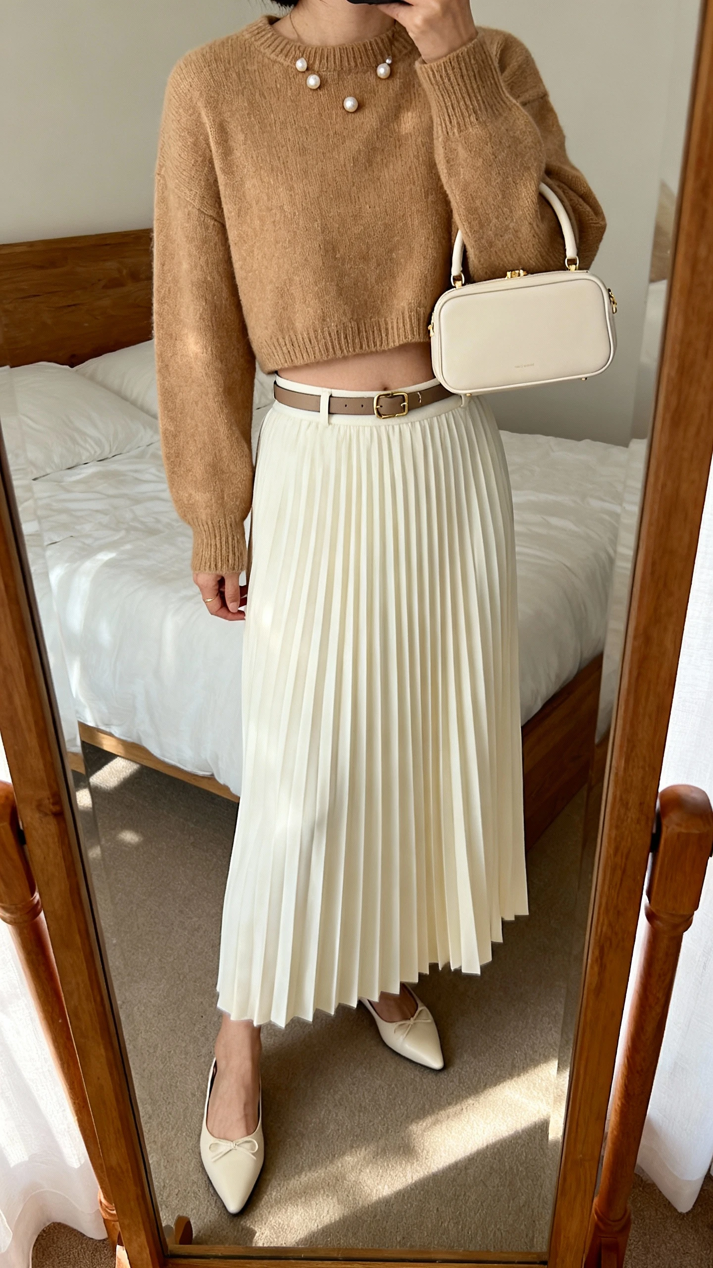 iPhone mirror selfie of a woman wearing a pleated midi skirt in cream with a cropped knit sweater in camel, pointed ballet flats, slim belt, pearl studs, and a compact top-handle bag, face not visible, cropped at shoulders, bedroom mirror, natural afternoon light, iPhone photo quality