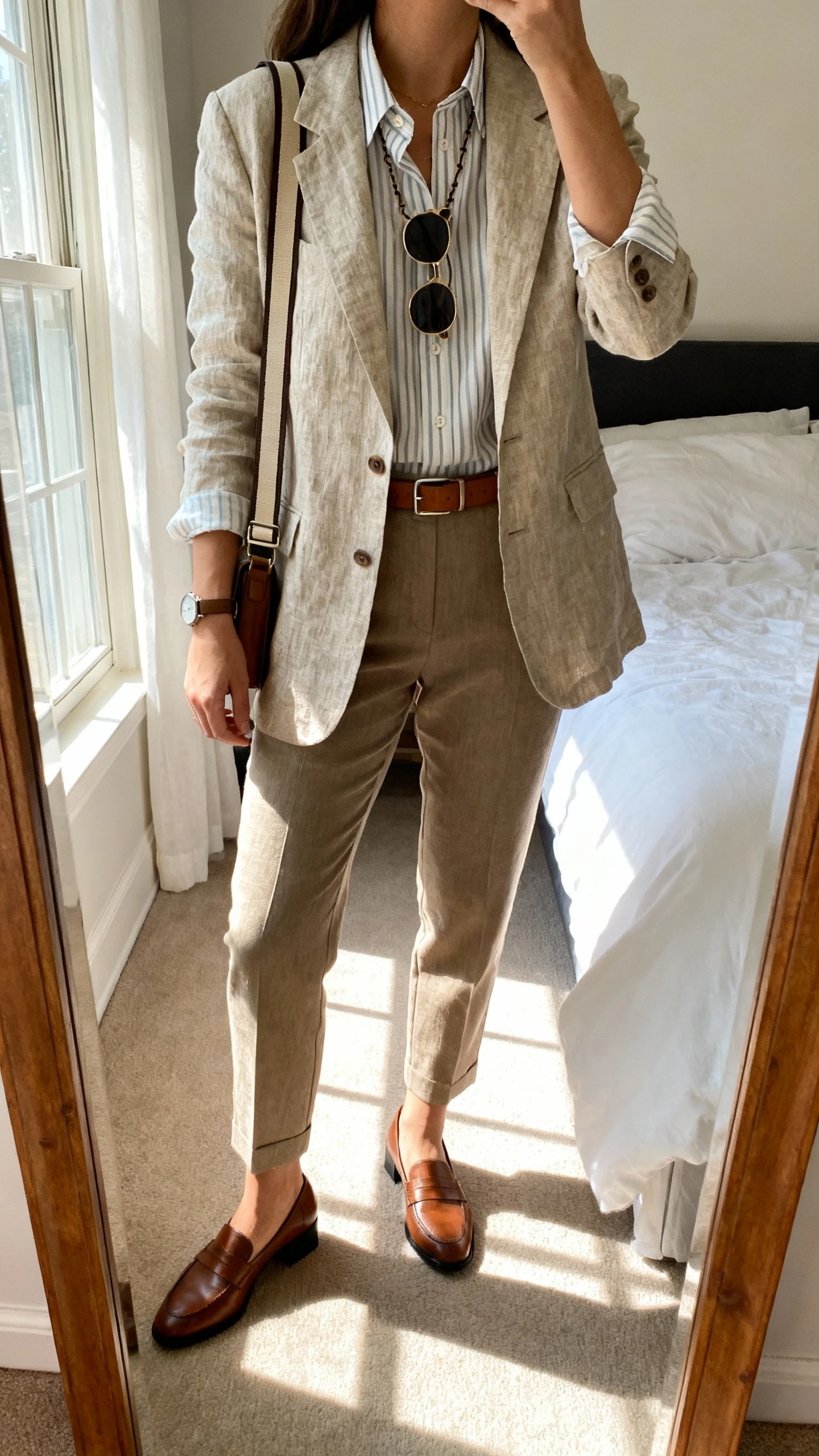 iPhone mirror selfie of a woman wearing a polished daywalker set: structured linen-blend blazer, breathable striped button-up, tailored ankle trousers, low block-heel leather loafers, crossbody with wide strap, slim leather belt, minimalist watch, and rubber-tipped sunglasses chain; face not visible, cropped at neck, bedroom full-length mirror with morning window light, iPhone photo quality.