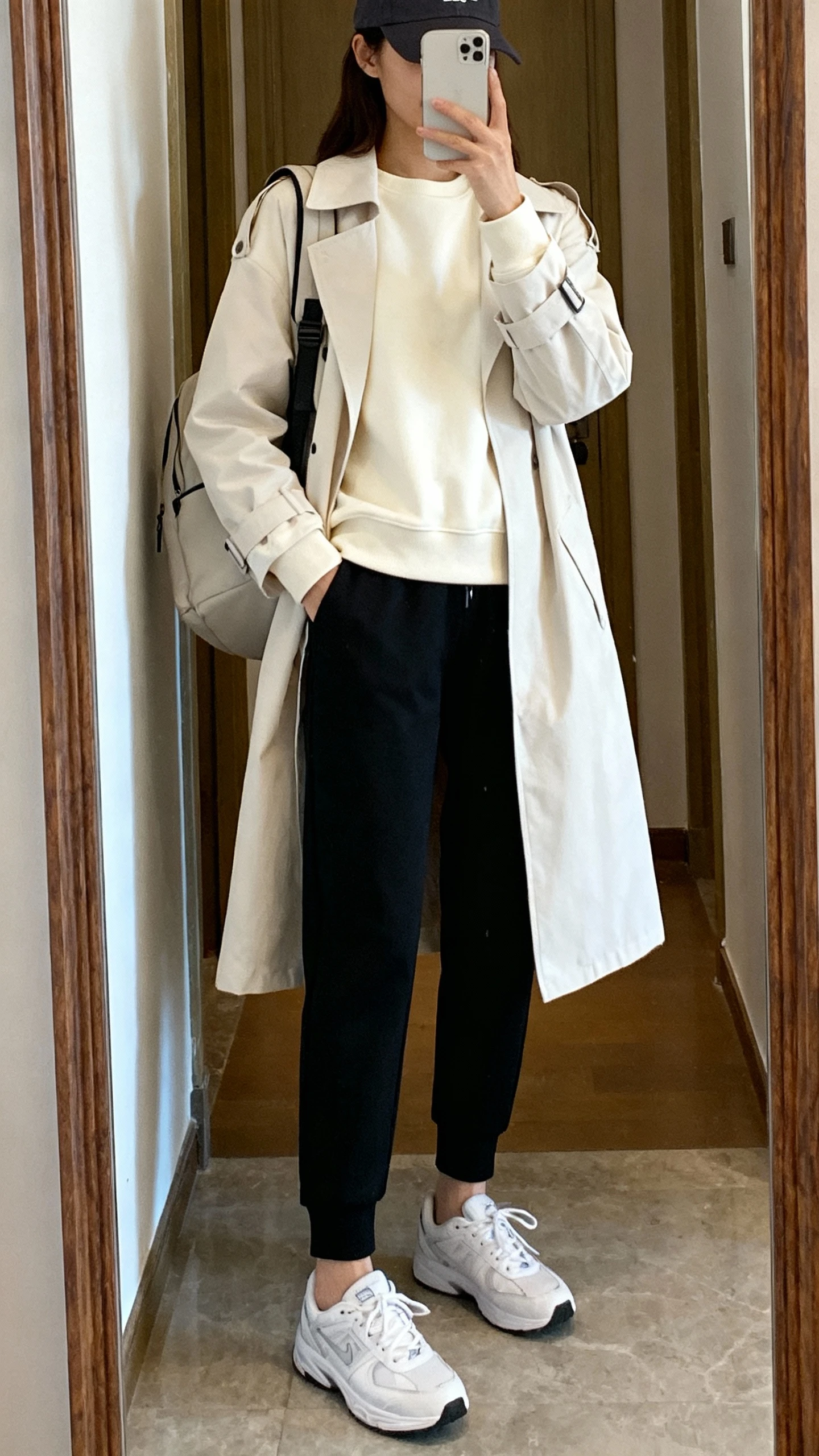 iPhone mirror selfie of a woman wearing a polished athleisure mix: black tapered joggers, a cream crewneck sweatshirt layered under a lightweight trench, sleek white sneakers, baseball cap, and a minimalist backpack, face not visible, cropped at neck, entryway mirror, natural daylight, iPhone photo quality.