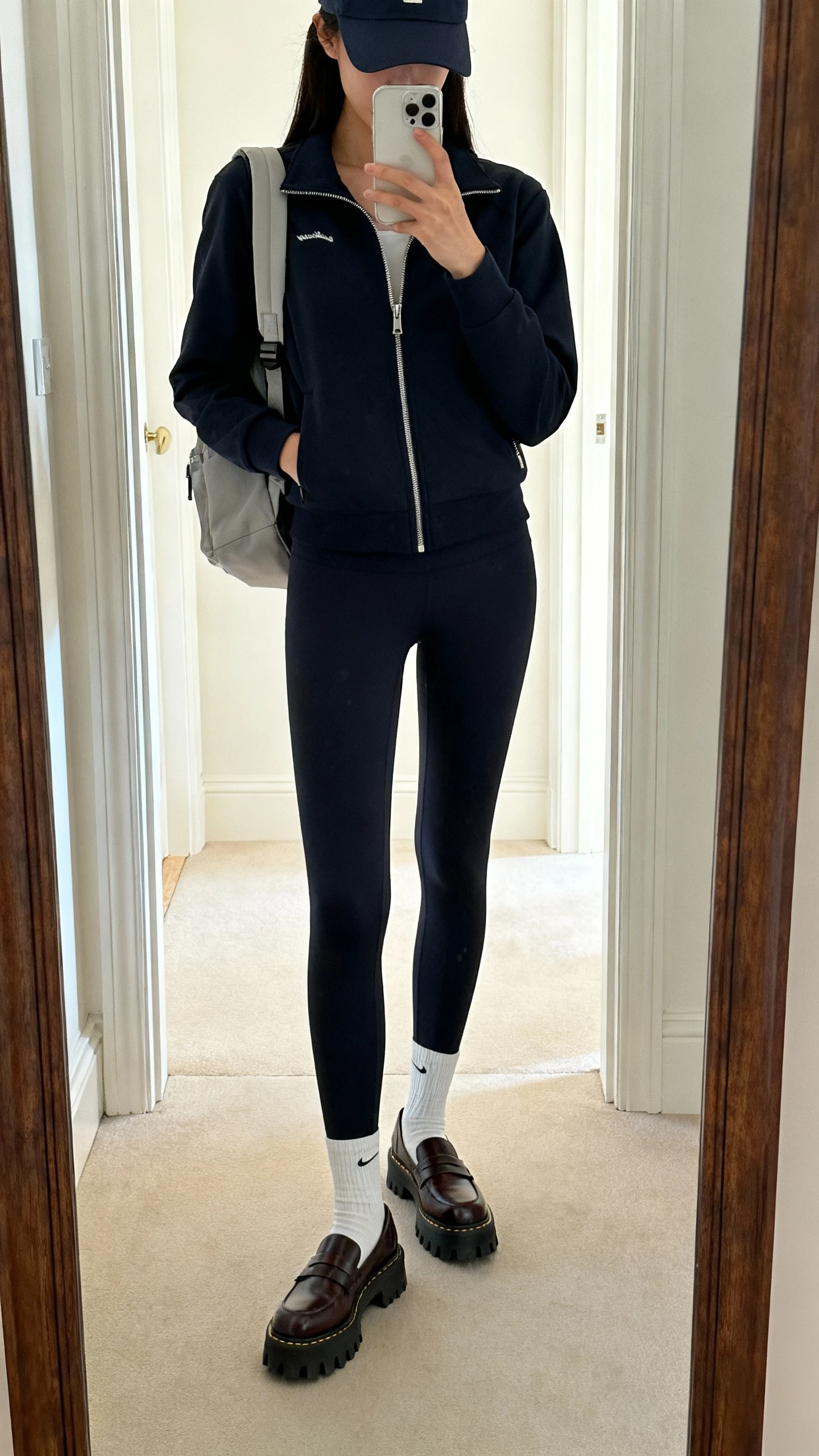 iPhone mirror selfie of a woman wearing a polished athleisure set (structured leggings and a zip-up track jacket), lug-sole boots or driving loafers with clean athletic socks, baseball cap and minimal backpack, face not visible, cropped at neck, entryway full-length mirror, bright indoor light, iPhone photo quality.