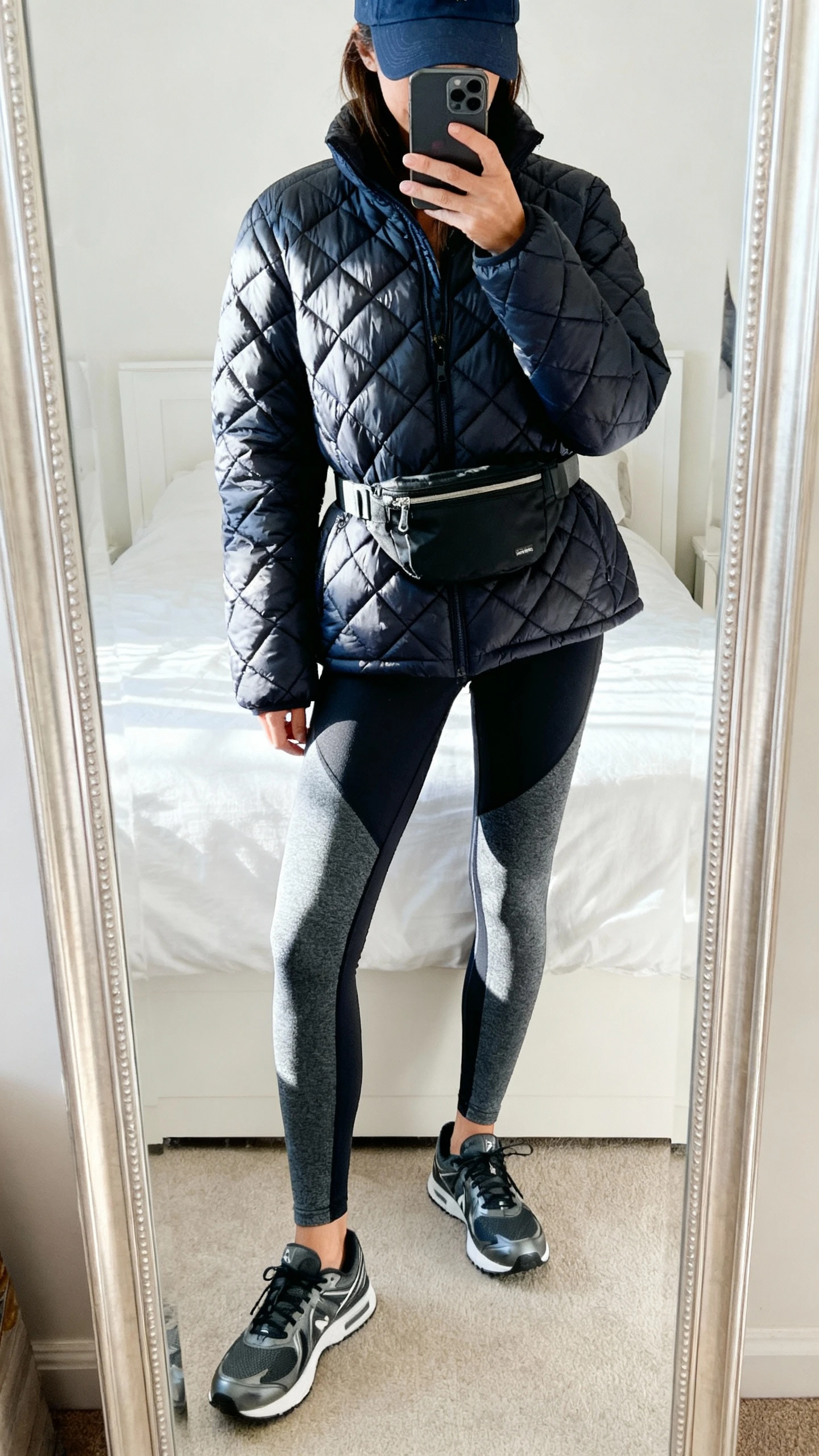 iPhone mirror selfie of a woman wearing a quilted puffer jacket, performance leggings, sleek trainers, baseball cap, and a belt bag, face not visible, cropped at shoulders, bedroom mirror, bright indoor lighting, iPhone photo quality.