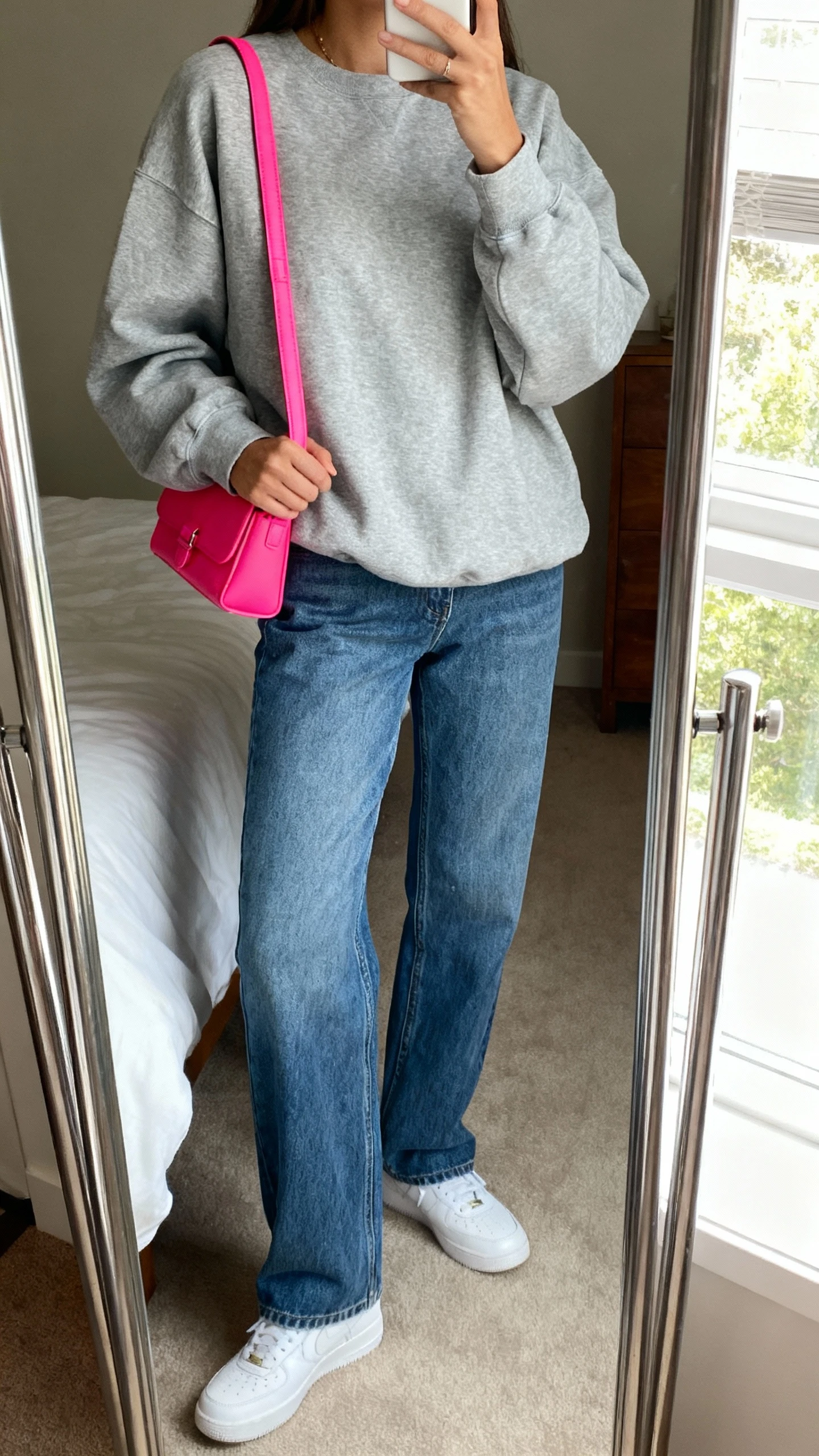iPhone mirror selfie of a woman wearing a relaxed gray sweatshirt, straight-leg blue jeans, white sneakers, and a bright pop-of-color shoulder bag, face not visible, cropped at neck, bedroom mirror, soft morning window light, iPhone photo quality.