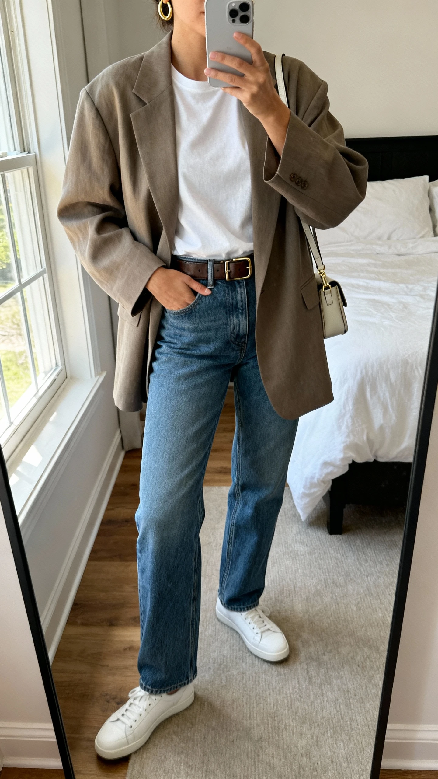 iPhone mirror selfie of a woman wearing a relaxed oversized blazer over a plain white tee, straight-leg blue jeans, sleek leather belt, minimalist white sneakers, small crossbody bag, and simple gold hoops, face not visible, cropped at neck, bedroom mirror with natural morning window light, iPhone photo quality.