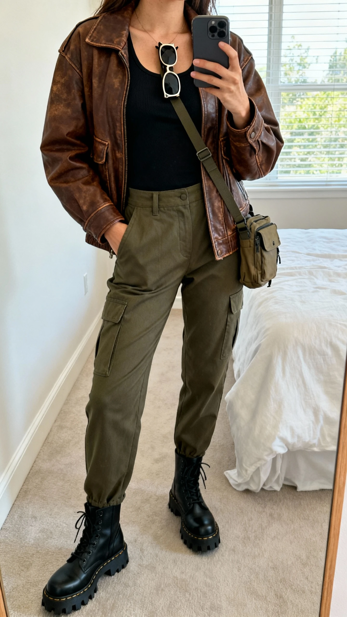 iPhone mirror selfie of a woman wearing a relaxed vintage-inspired brown leather jacket, fitted black tank, tailored olive cargo pants, black lug-sole boots, rectangular sunglasses tucked on neckline, and a crossbody utility bag; face not visible, cropped at shoulders, bedroom mirror, daytime window light, iPhone photo quality.