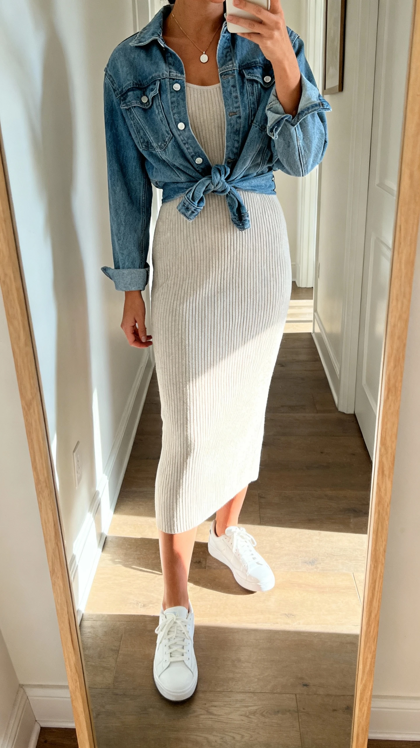 iPhone mirror selfie of a woman wearing a ribbed knit midi dress with white sneakers, denim jacket tied at the waist, subtle pendant necklace, face not visible, cropped at shoulders, hallway full-length mirror, bright morning light, iPhone photo quality