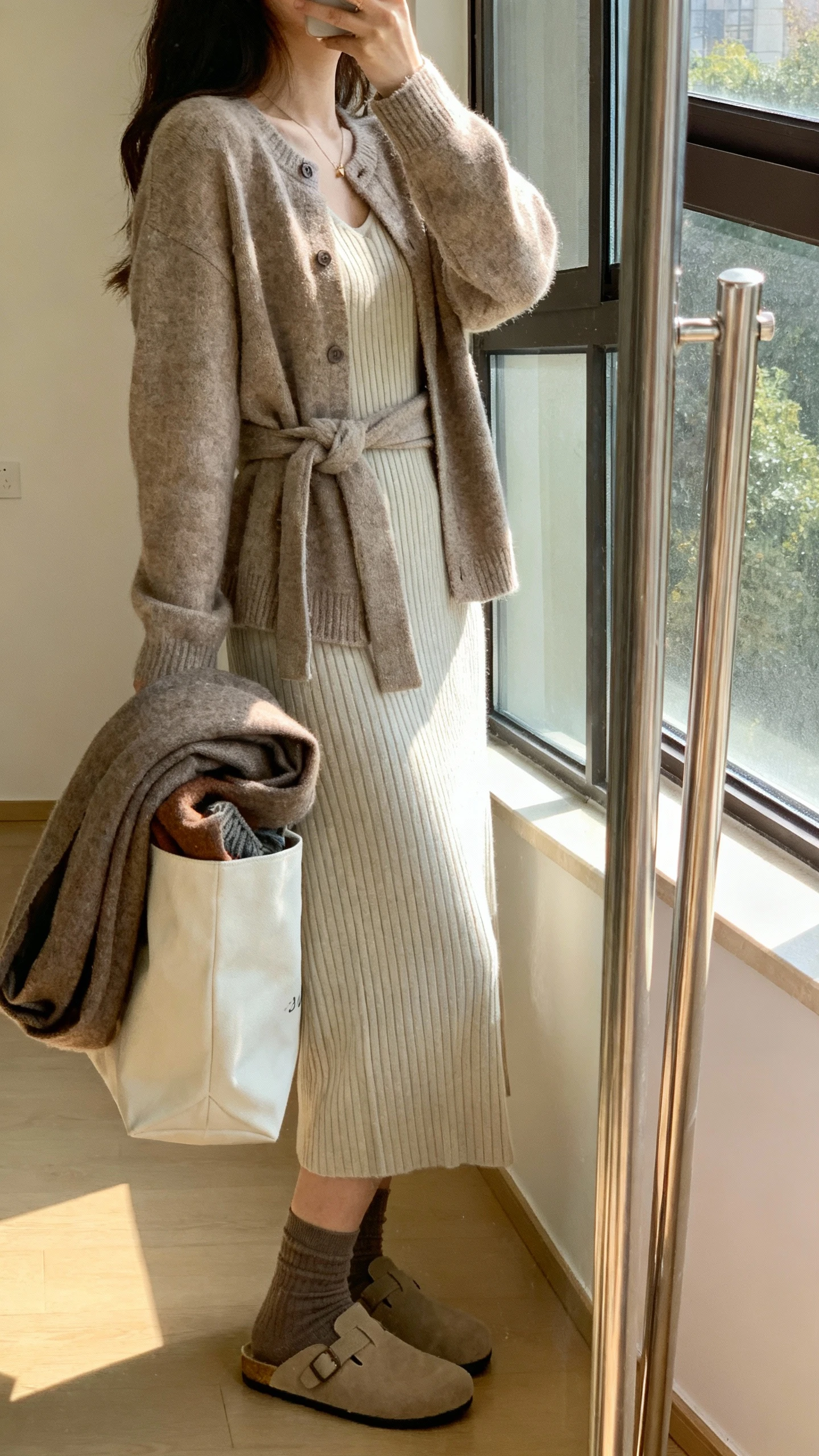 iPhone mirror selfie of a woman wearing a ribbed midi dress with a cozy cardigan tied at the waist, slip-on flats, oversized scarf tucked in tote, and ankle socks, face not visible, side angle, full-length mirror by window, gentle morning light, iPhone photo quality.