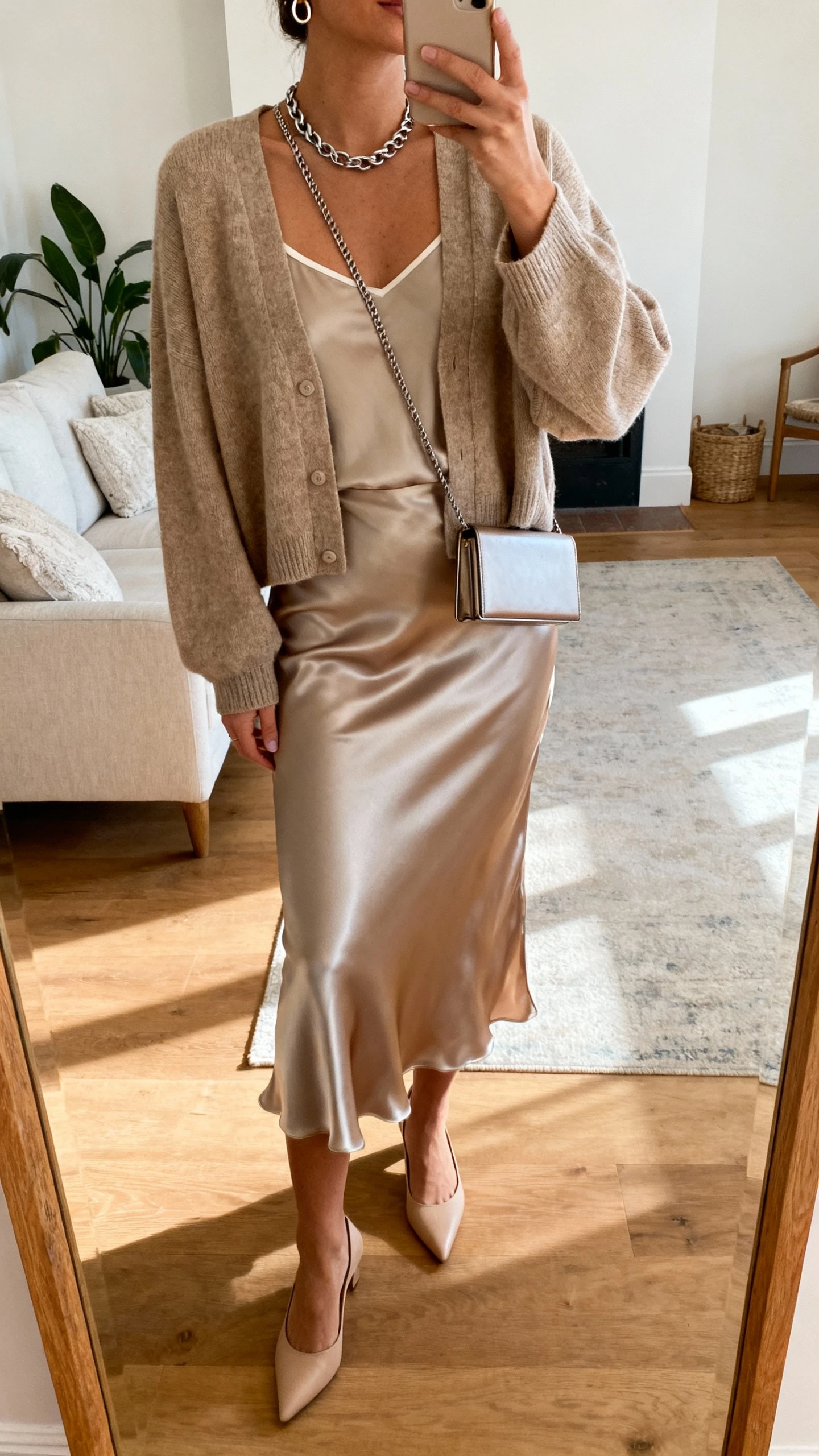 iPhone mirror selfie of a woman wearing a satin bias-cut midi slip skirt, a boxy cardigan over a thin camisole, minimalist kitten heels, a chain necklace, small hoops, and a slim clutch-crossbody; face not visible, cropped at neck, living room full-length mirror, warm indoor lighting, iPhone photo quality.