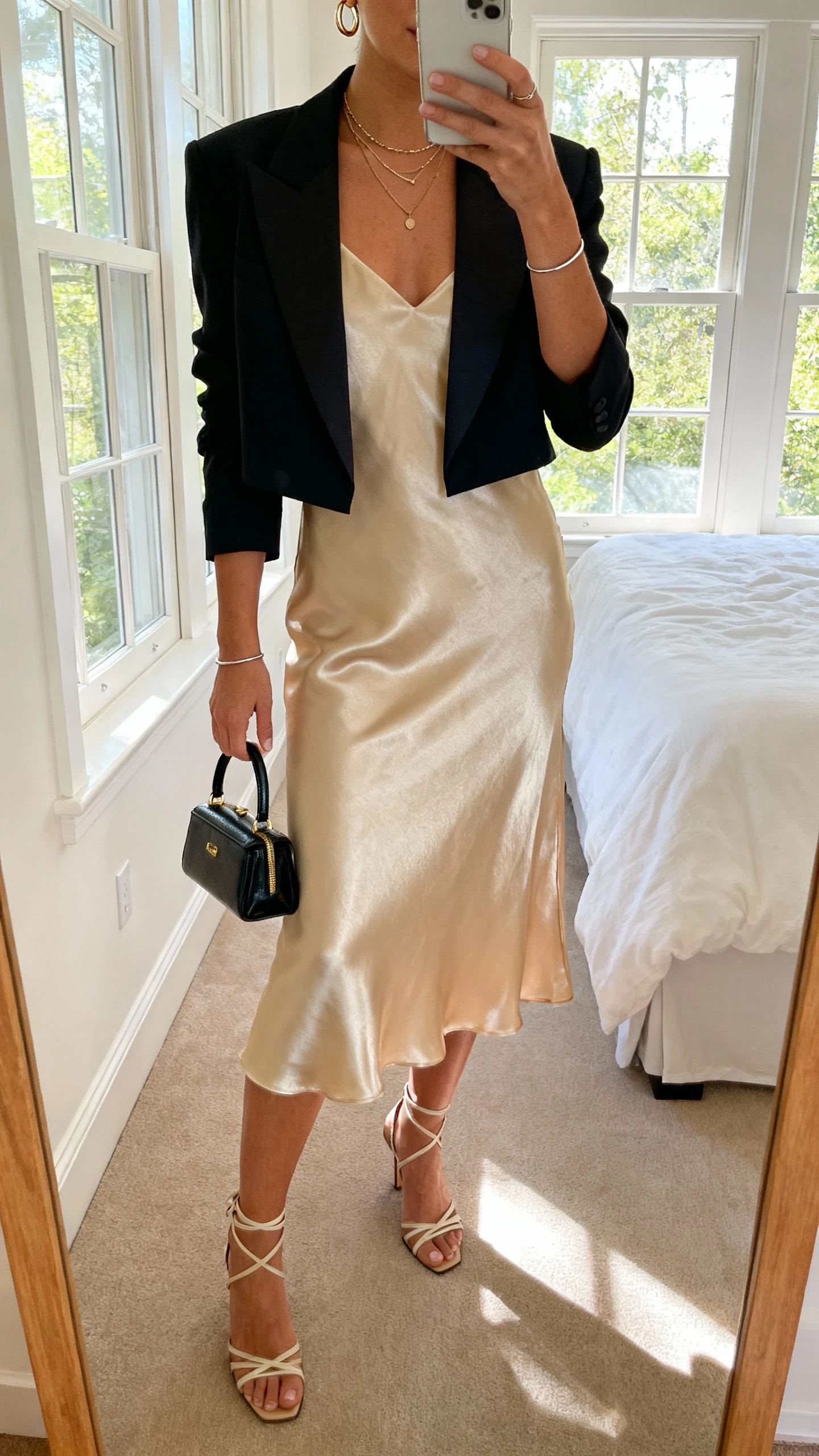 iPhone mirror selfie of a woman wearing a satin slip dress in champagne with a cropped tuxedo blazer, strappy heels, a mini top-handle bag, delicate layered necklaces, huggie hoops, and a thin bracelet; face not visible, cropped at neck, bedroom mirror with natural window light, iPhone photo quality.