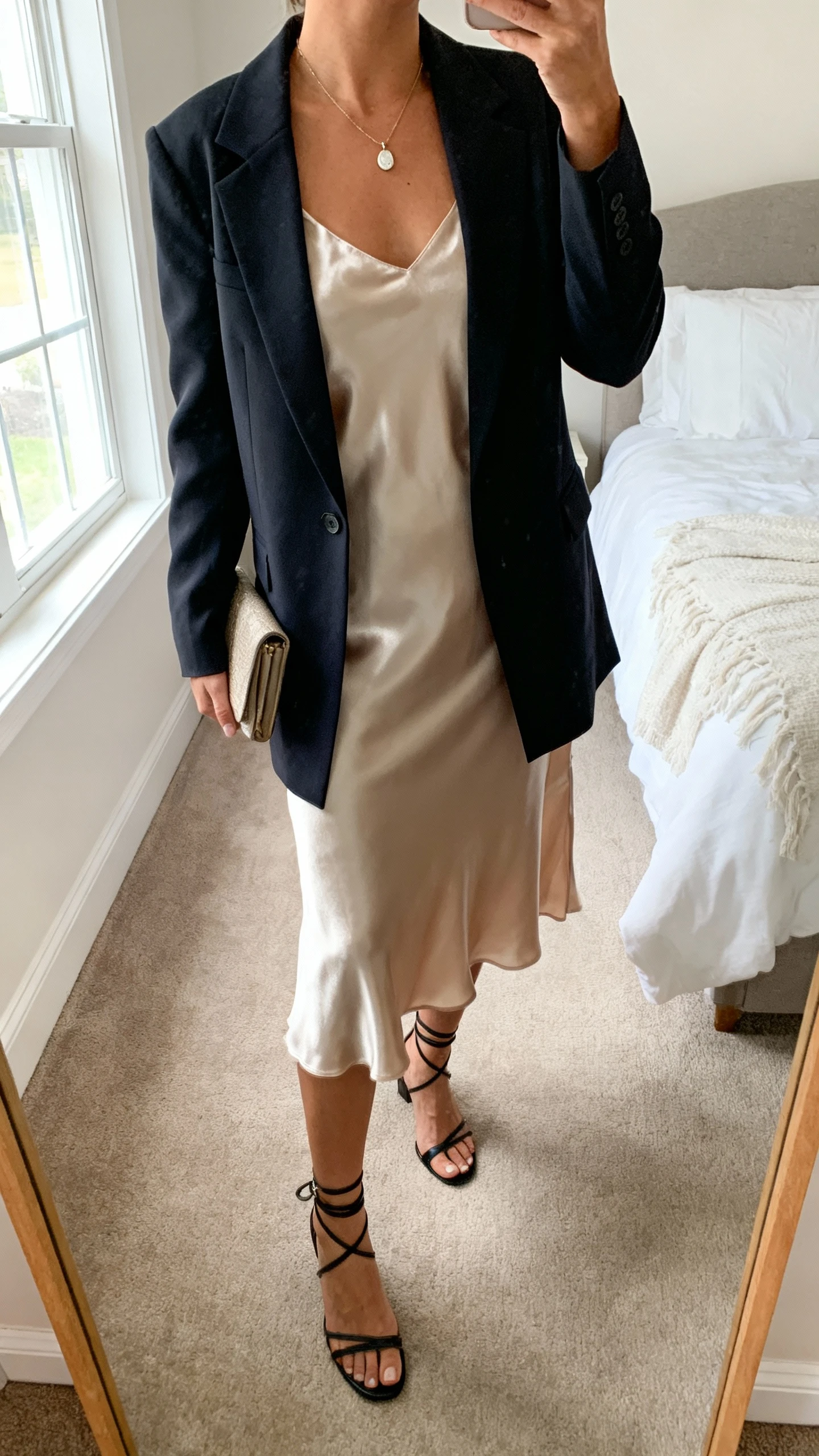 iPhone mirror selfie of a woman wearing a satin slip dress layered under a tailored blazer, delicate pendant necklace, small clutch, and strappy heels; face not visible, cropped at neck, bedroom mirror with natural window light, iPhone photo quality.