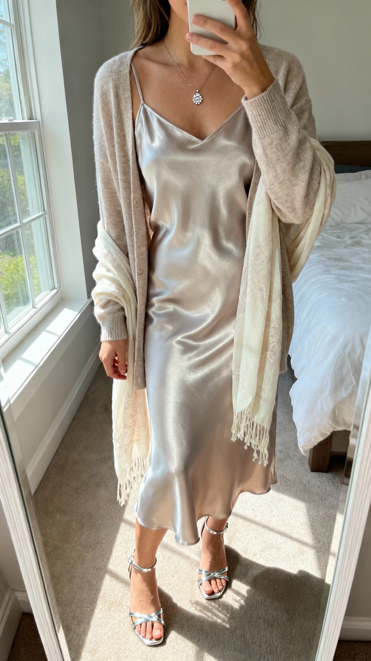 iPhone mirror selfie of a woman wearing a satin slip dress layered with a soft cardigan and delicate shawl, minimal pendant necklace and strappy heels, face not visible, cropped at neck, bedroom mirror with natural window light, iPhone photo quality