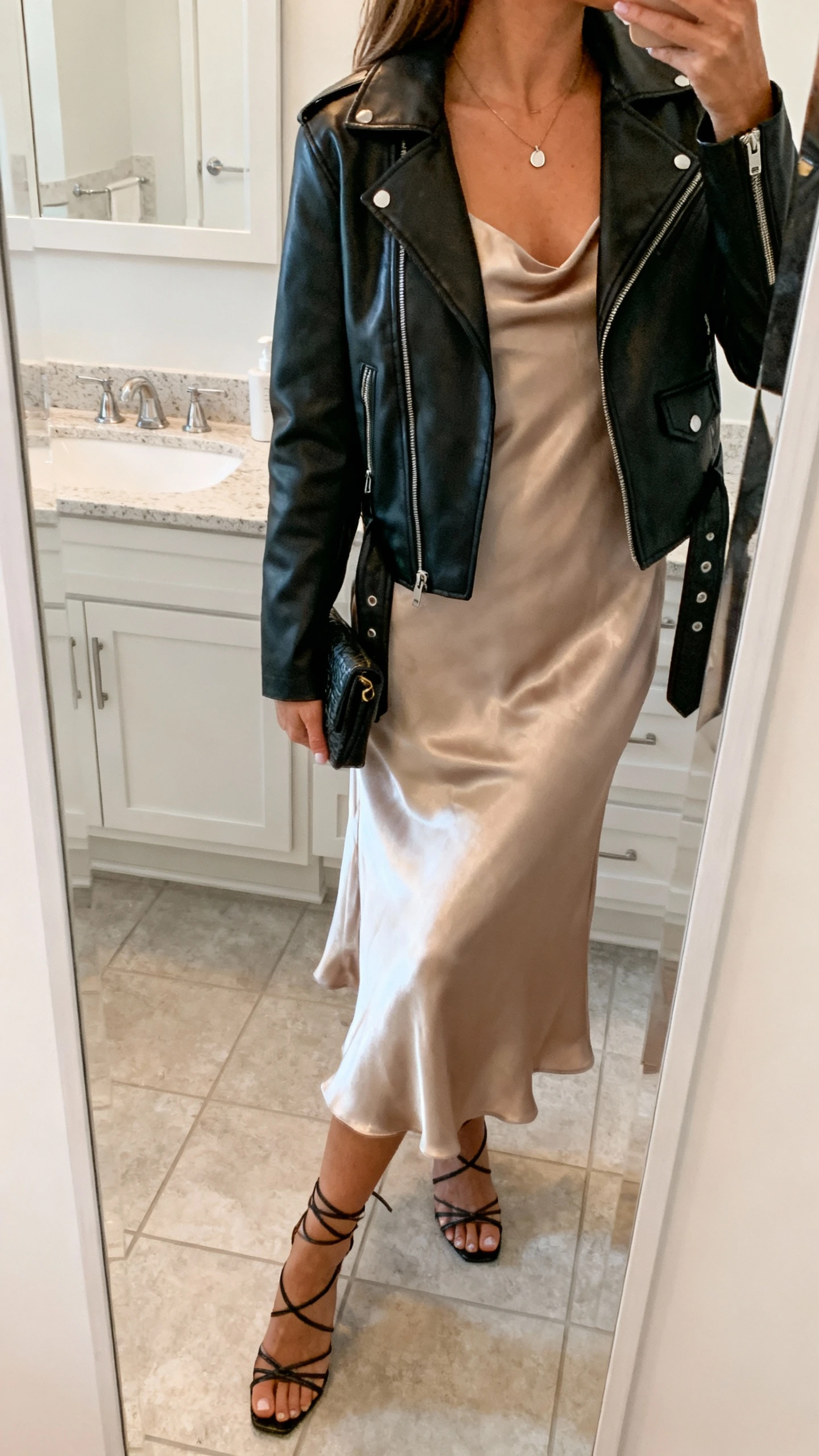 iPhone mirror selfie of a woman wearing a satin slip dress layered with a black leather moto jacket, minimal pendant necklace, strappy heels, and a small clutch, face not visible, side angle in a bathroom full-length mirror with soft indoor lighting, iPhone photo quality.