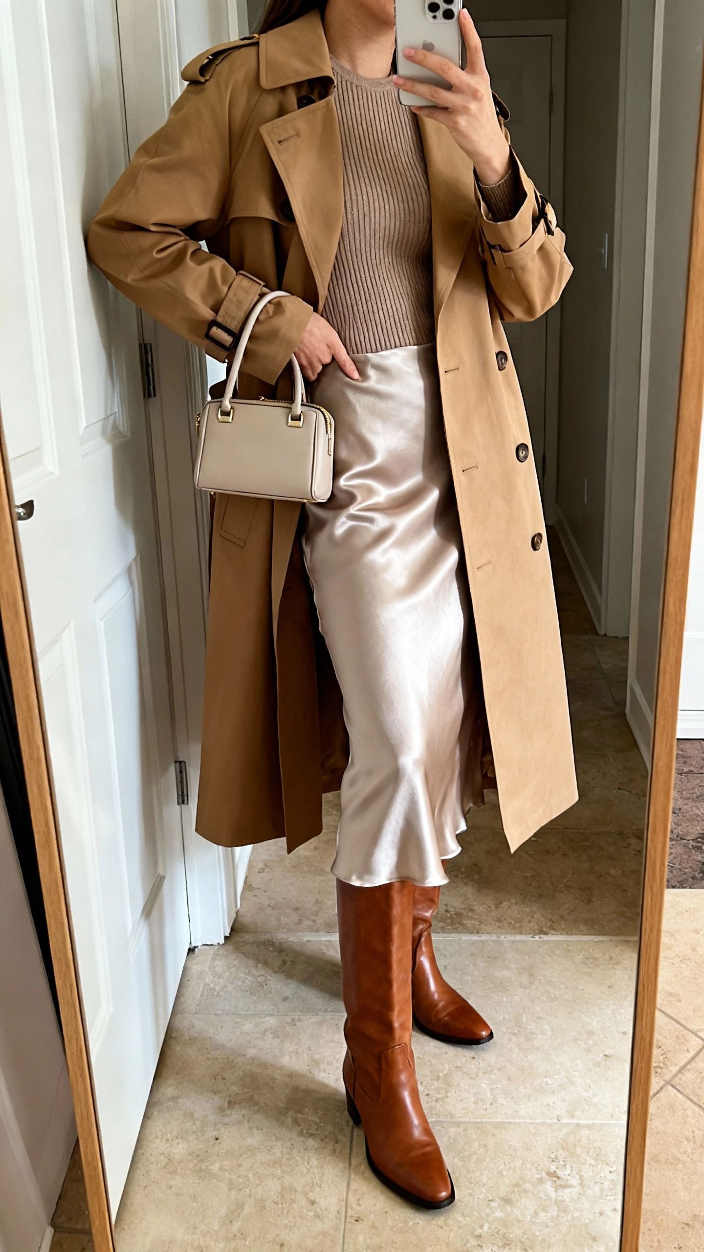 iPhone mirror selfie of a woman wearing a satin slip skirt, classic camel trench coat over a fitted knit top, mid-calf leather boots, and a small top-handle bag, face not visible, side angle, entryway full-length mirror, natural daylight, iPhone photo quality.