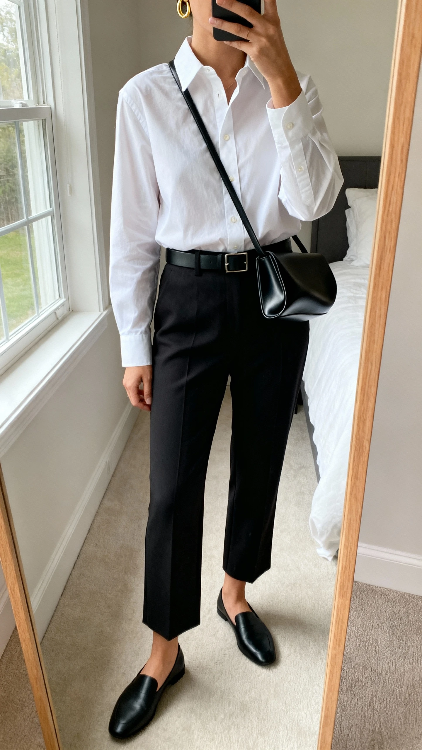 iPhone mirror selfie of a woman wearing a sculptural black crossbody bag, crisp white button-up shirt tucked into tailored black cigarette trousers, slim black belt, minimalist black loafers, and a fine gold hoop earring peeking in frame; face not visible, cropped at neck, bedroom mirror, natural window light, iPhone photo quality.