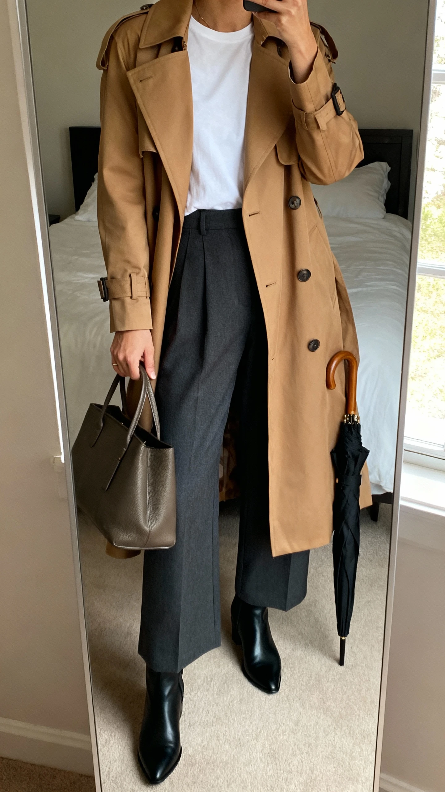 iPhone mirror selfie of a woman wearing a sharp camel trench coat over a crisp white tee, charcoal wide-leg trousers, sleek black ankle boots, structured leather tote, and a compact black umbrella in hand, face not visible, cropped at neck, bedroom mirror with natural window light, iPhone photo quality.