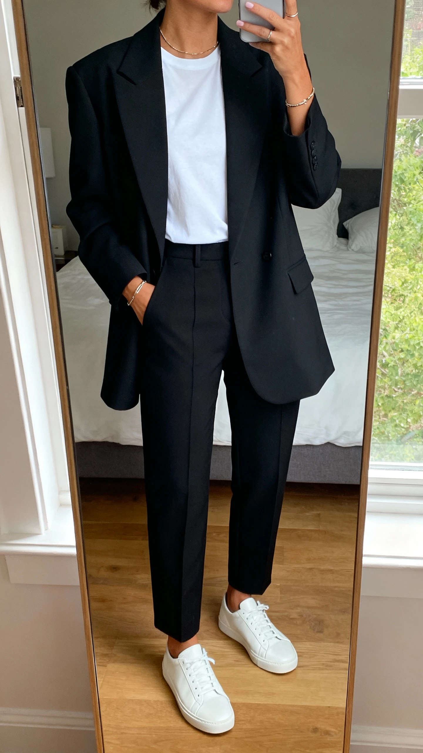 iPhone mirror selfie of a woman wearing a sharp oversized power blazer over a crisp white tee, tailored black trousers, minimalist jewelry, and clean white leather sneakers, face not visible, cropped at neck, bedroom mirror, natural window light, iPhone photo quality.