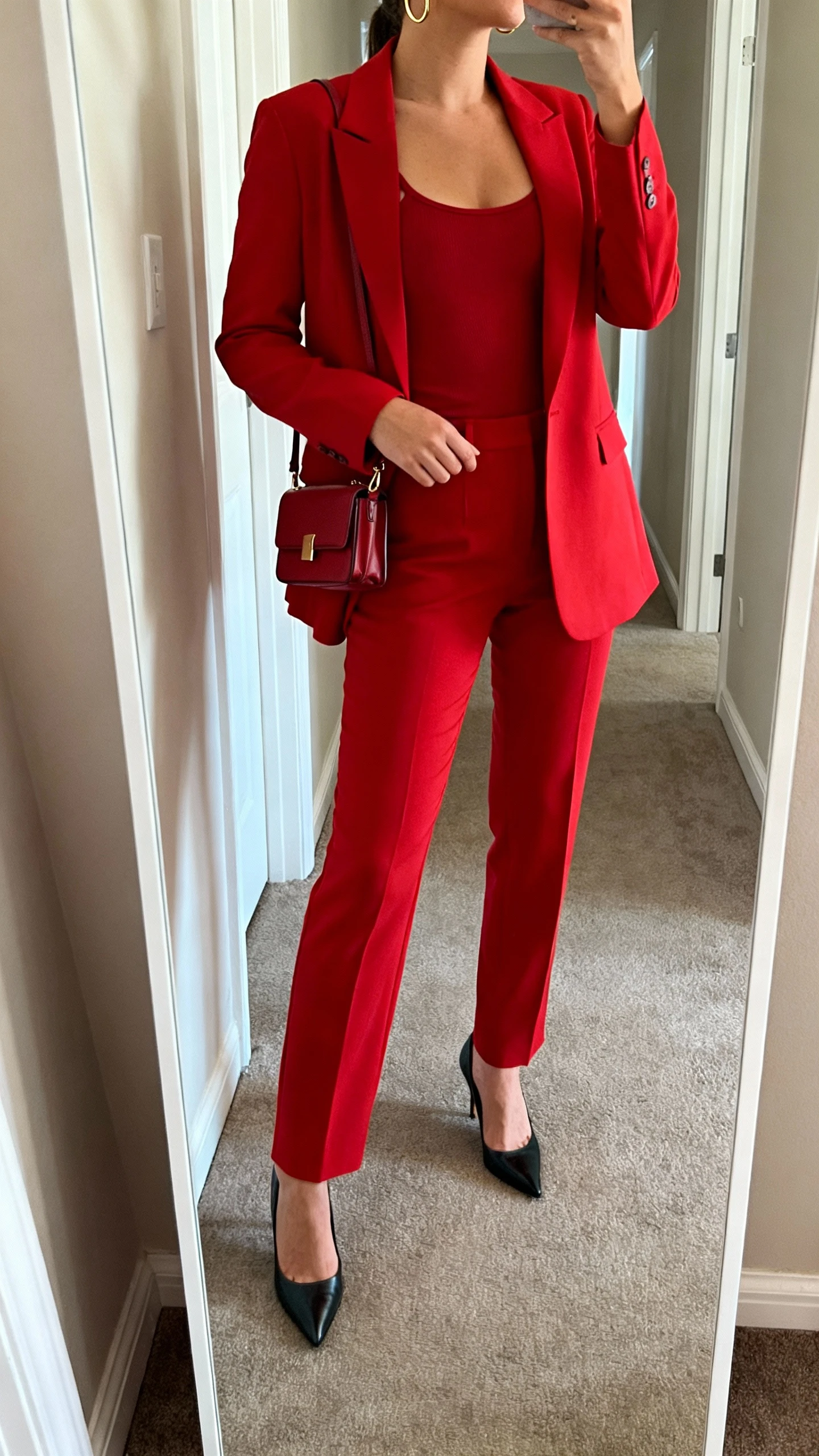iPhone mirror selfie of a woman wearing a sharp red suit (blazer and tailored trousers) with a fitted camisole, pointed heels, structured mini bag, and minimal gold hoops, face not visible, cropped at shoulders, full-length hallway mirror, natural daylight, iPhone photo quality.