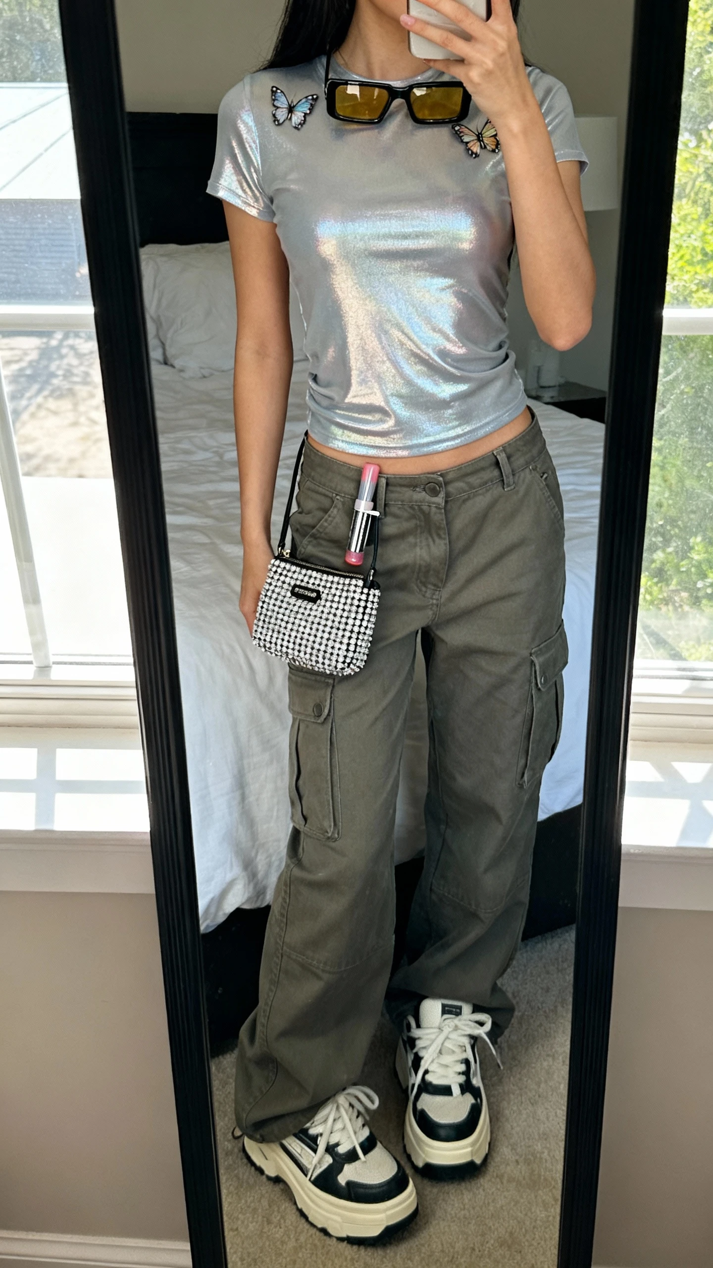 iPhone mirror selfie of a woman wearing a shiny metallic baby tee, low-rise cargo pants, chunky platform sneakers, a rhinestone-studded mini bag, tinted rectangle sunglasses perched on head, glossy lip roller clipped to belt loop, and butterfly hair clips, face not visible, cropped at neck, bedroom mirror, natural window light, iPhone photo quality
