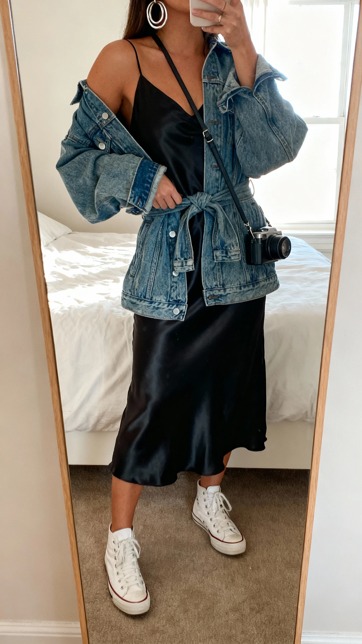 iPhone mirror selfie of a woman wearing a silky black midi slip dress with white retro sneakers, oversized denim jacket tied at the waist, chunky silver hoops, slim crossbody camera bag, face not visible, cropped at shoulders, bedroom mirror, bright window light, iPhone photo quality.