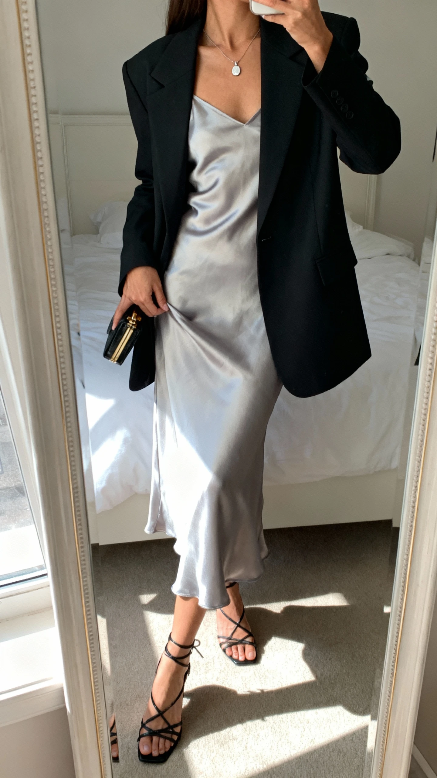 iPhone mirror selfie of a woman wearing a silky slip dress with a sharp tailored blazer, delicate pendant necklace, strappy heels, and a small clutch in hand, face not visible, cropped at neck, bedroom mirror, soft morning window light, iPhone photo quality.