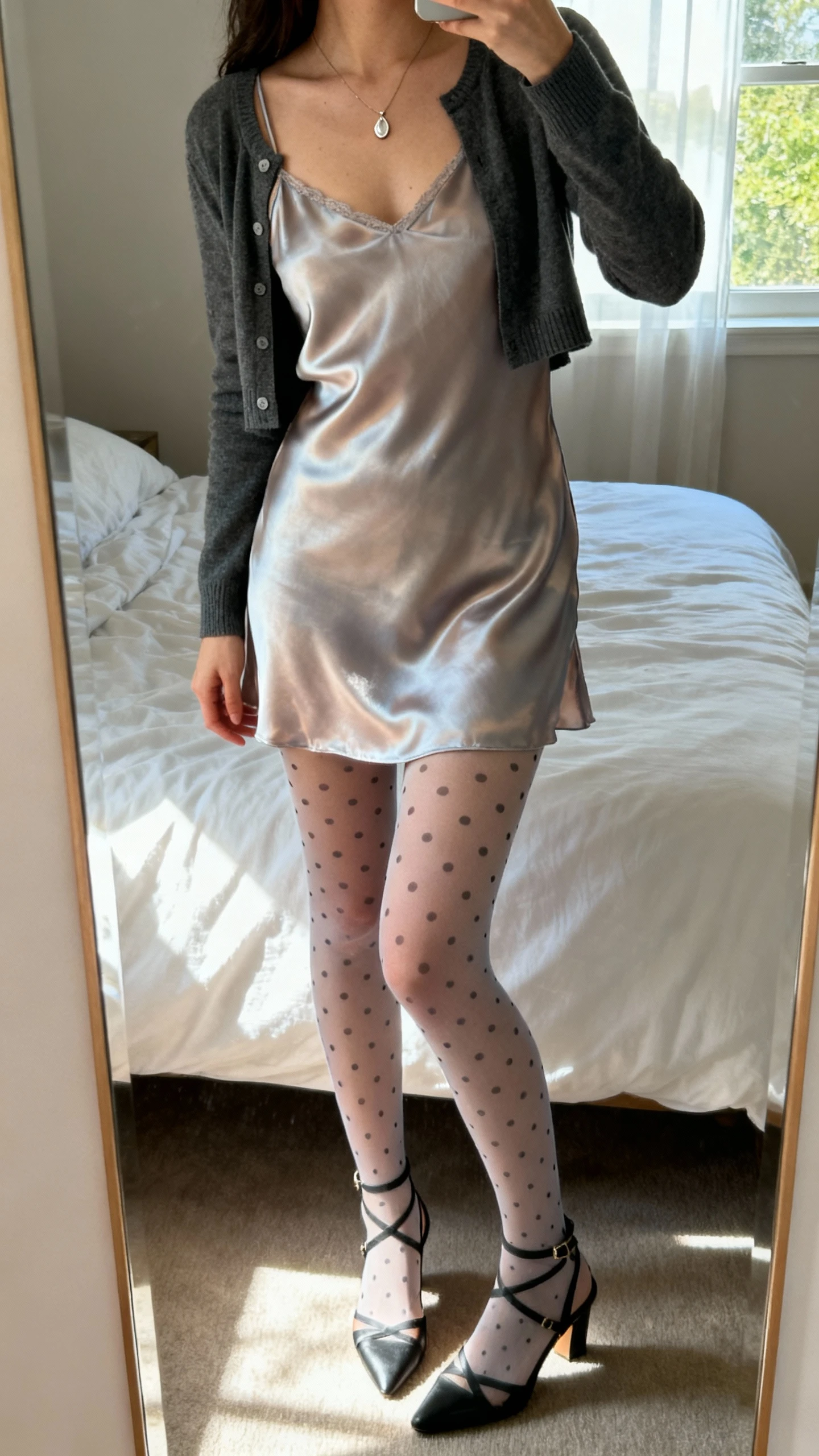 iPhone mirror selfie of a woman wearing a silky slip dress layered over patterned stockings (subtle polka dots), strappy heels, cropped cardigan, and delicate pendant necklace, face not visible, cropped at shoulders, bedroom mirror with natural window light, iPhone photo quality.