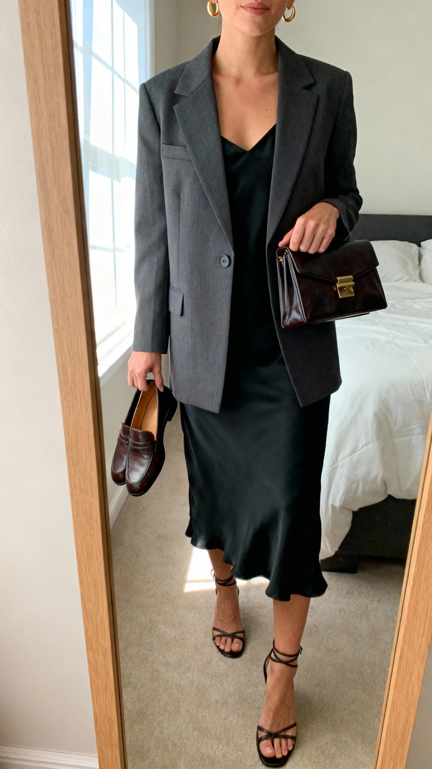 iPhone mirror selfie of a woman wearing a single-breasted charcoal tailored blazer over a black bias-cut silk midi slip dress, with leather loafers in hand and strappy heels on, small gold hoops and a structured leather clutch; face not visible, cropped at neck, bedroom mirror, natural window light, iPhone photo quality.