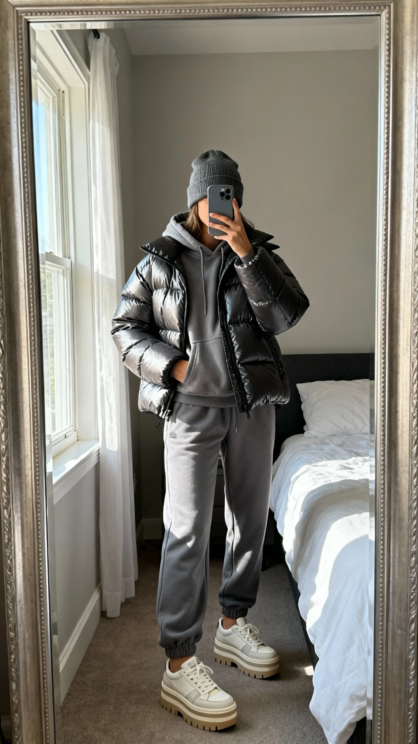 iPhone mirror selfie of a woman wearing a sleek puffer jacket over a matching sweats set (hoodie + joggers) with platform sneakers and a beanie, face not visible, full-length bedroom mirror, natural window light, iPhone photo quality.