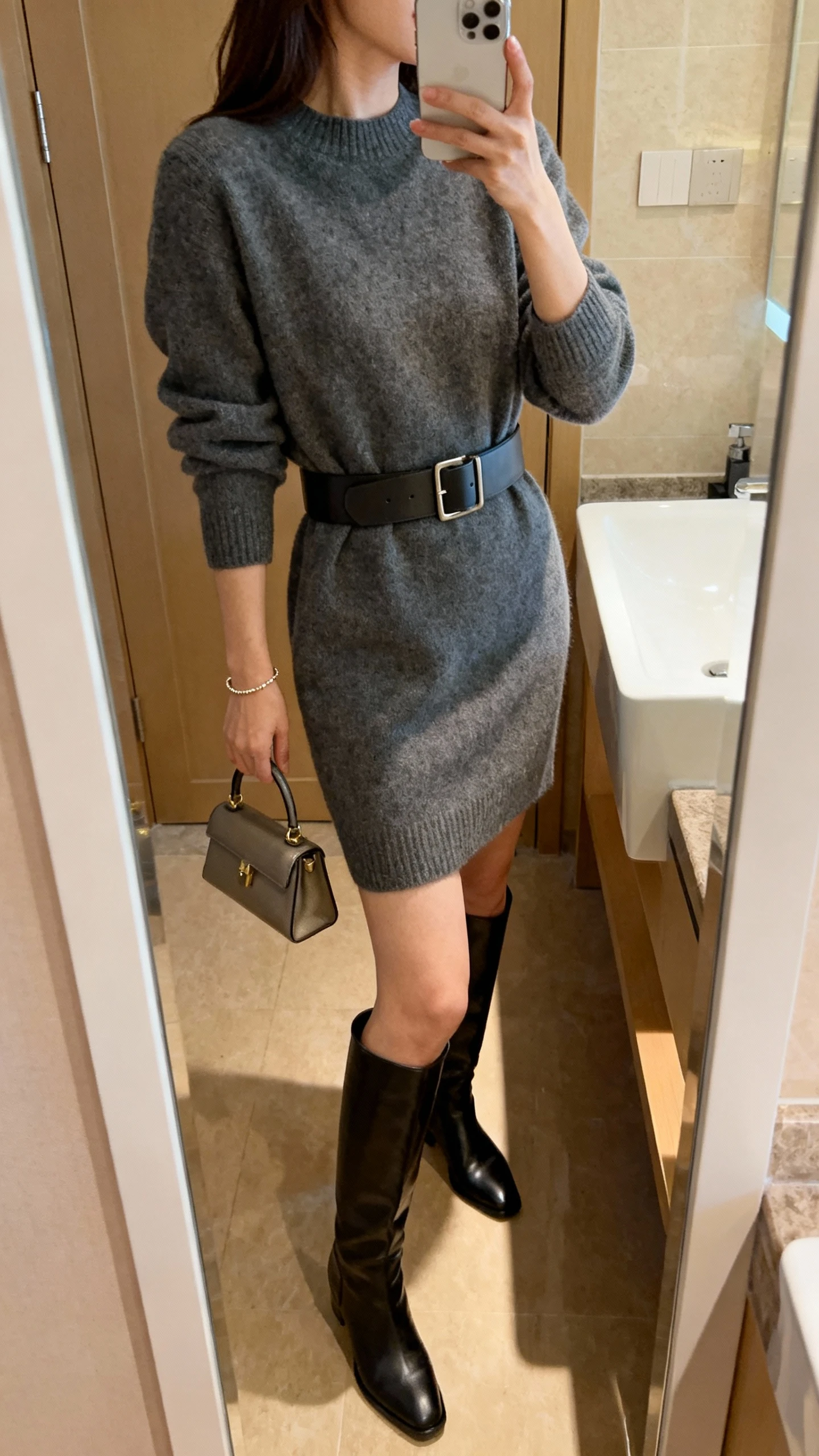 iPhone mirror selfie of a woman wearing a sleek sweater dress cinched with a structured belt and over-the-knee boots, face not visible, side angle in a bathroom full-length mirror, indoor warm lighting, iPhone photo quality; small top-handle bag and delicate bracelet.