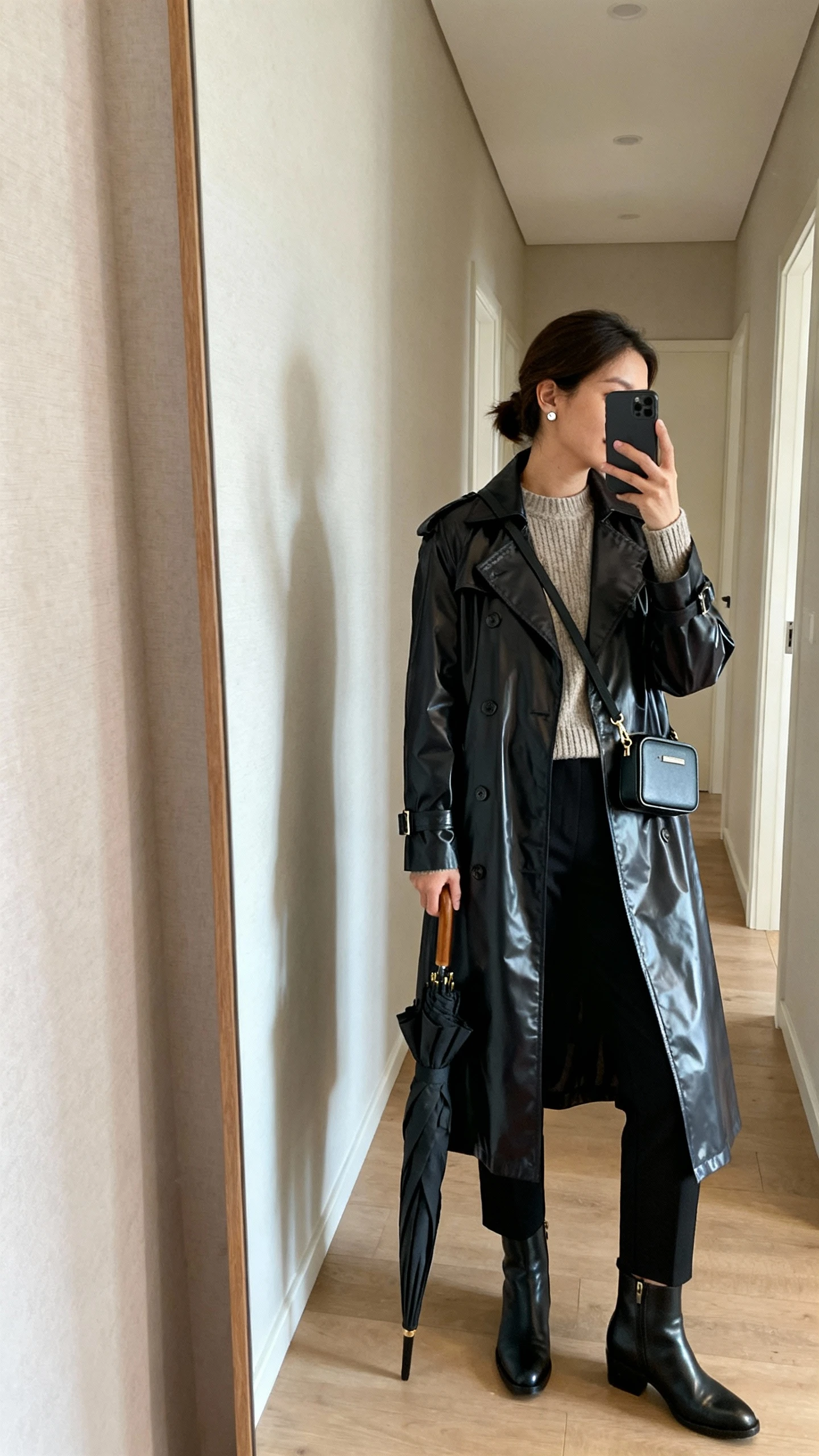 iPhone mirror selfie of a woman wearing a sleek waterproof trench, fine-knit crewneck, tapered black trousers, leather ankle boots, compact crossbody bag, and a folded umbrella in hand with subtle stud earrings, face not visible, side angle, hallway full-length mirror with soft indoor light, iPhone photo quality