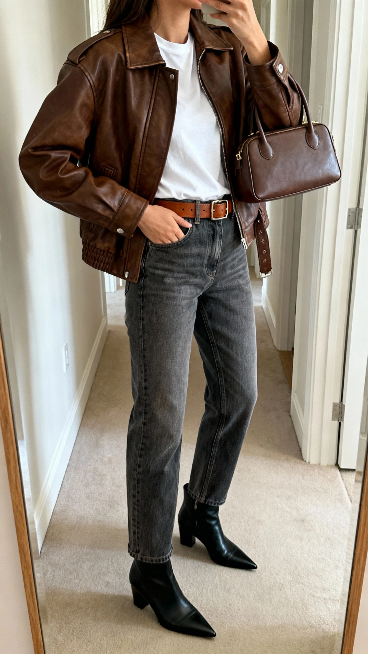 iPhone mirror selfie of a woman wearing a slightly oversized brown leather biker jacket, crisp white tee, medium-wash straight-leg jeans, pointed-toe black ankle boots, coordinating brown leather belt, and a structured top-handle bag; face not visible, side angle in hallway full-length mirror, soft indoor light, iPhone photo quality.