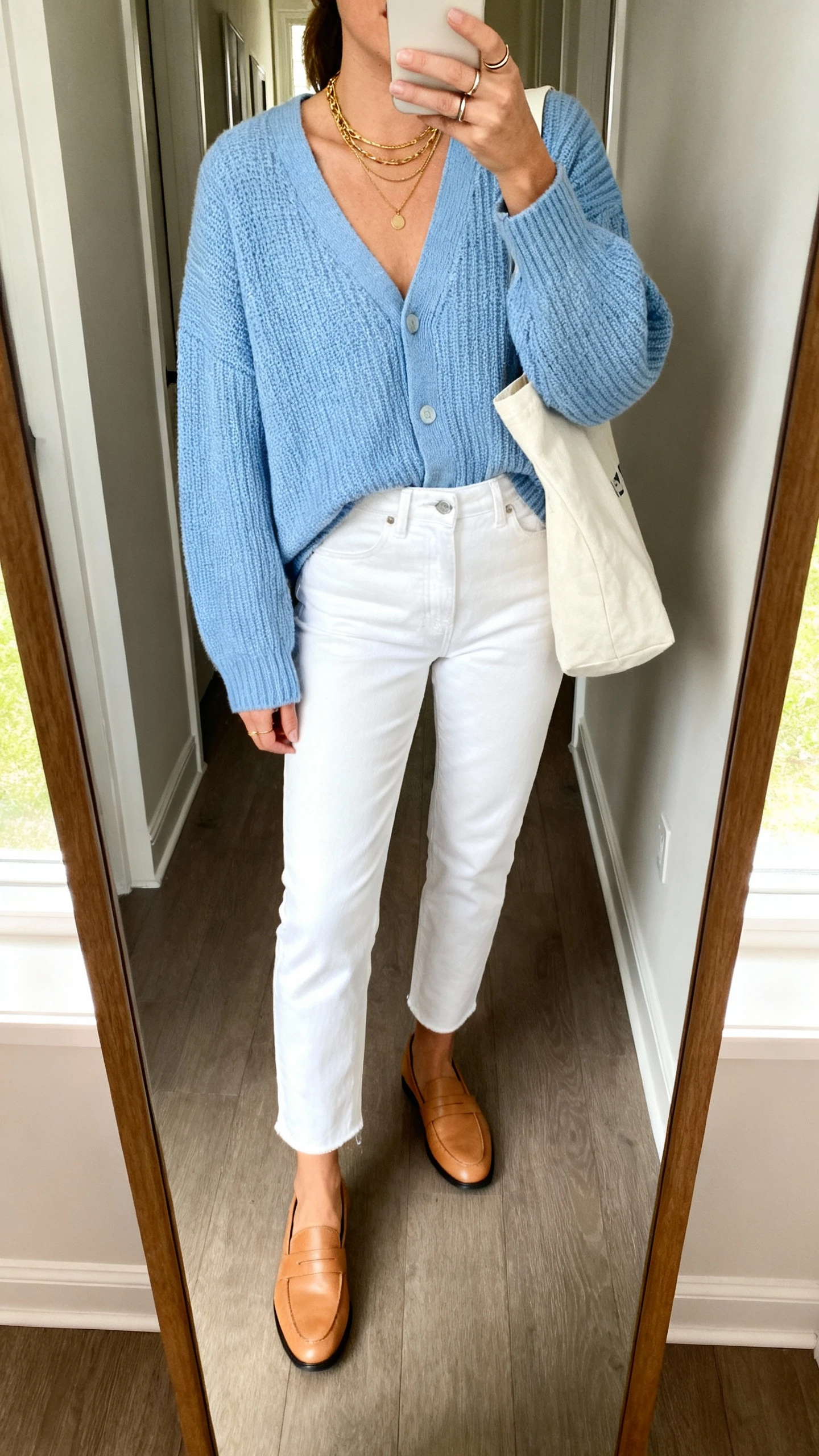 iPhone mirror selfie of a woman wearing a slightly oversized sky-blue fine-knit cardigan, high-rise straight-leg white ankle jeans, tan leather loafers, a cream canvas tote, layered gold necklaces and slim stacking rings, face not visible, cropped at shoulders, full-length hallway mirror, indoor daylight, iPhone photo quality.