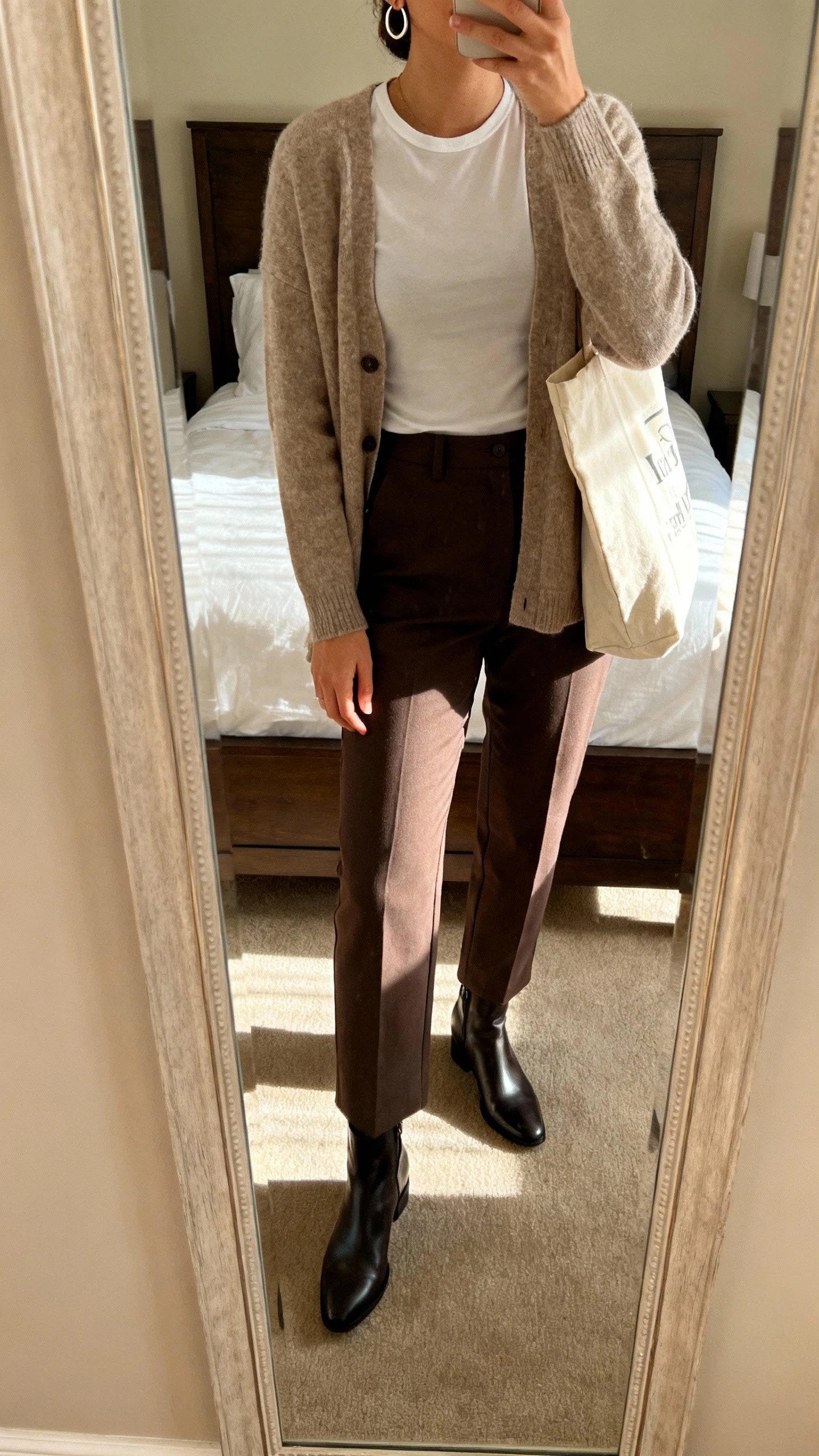 iPhone mirror selfie of a woman wearing a soft cardigan over a fitted tee, straight-leg trousers, sleek ankle boots, simple hoop earrings, canvas tote, face not visible, cropped at neck, bedroom mirror, warm indoor morning light, iPhone photo quality