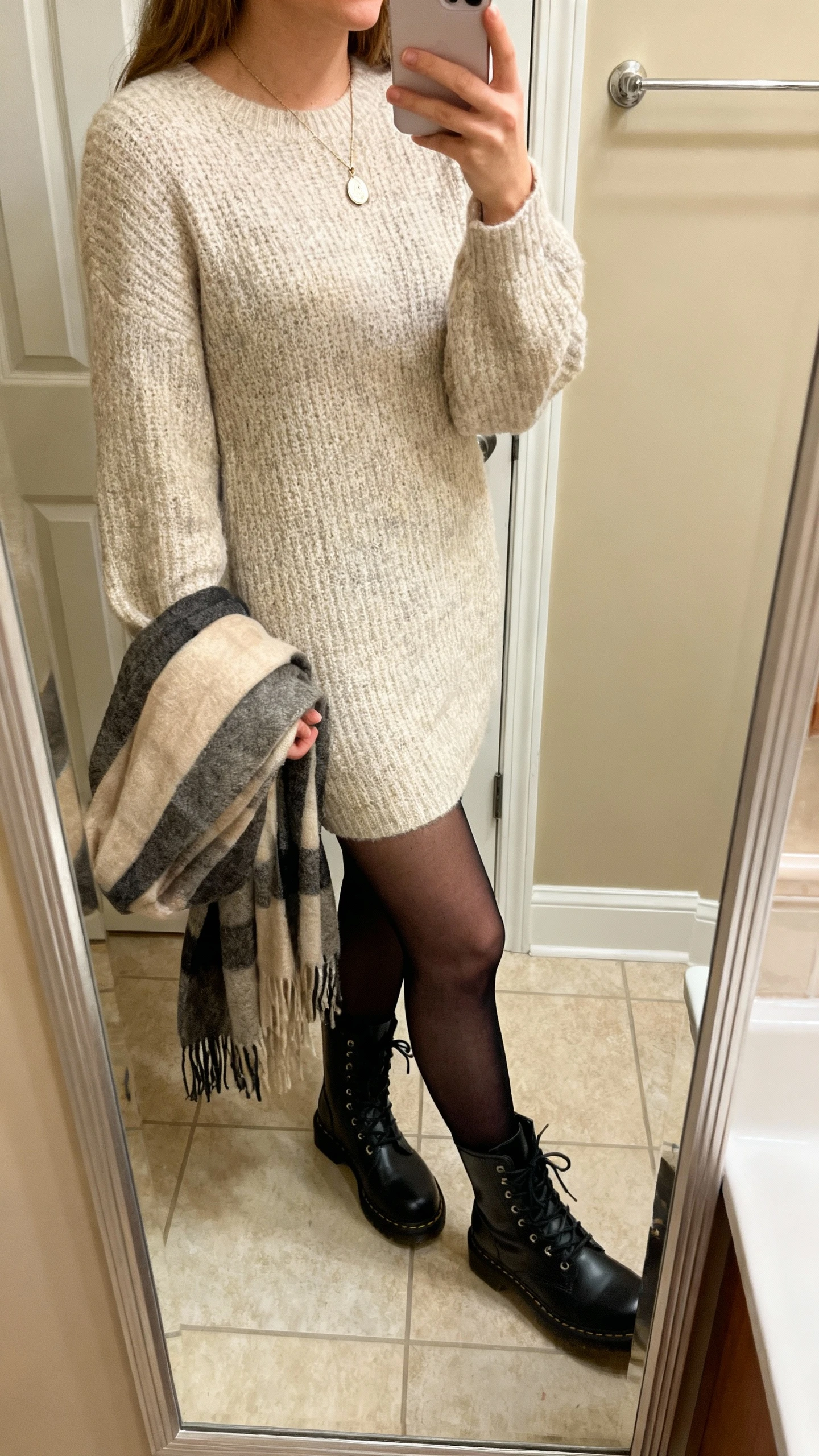 iPhone mirror selfie of a woman wearing a soft knit sweater dress, semi-opaque black tights, lace-up combat boots, delicate pendant necklace, and a cozy scarf draped over arm, face not visible, side angle, bathroom mirror, warm indoor lighting, iPhone photo quality.