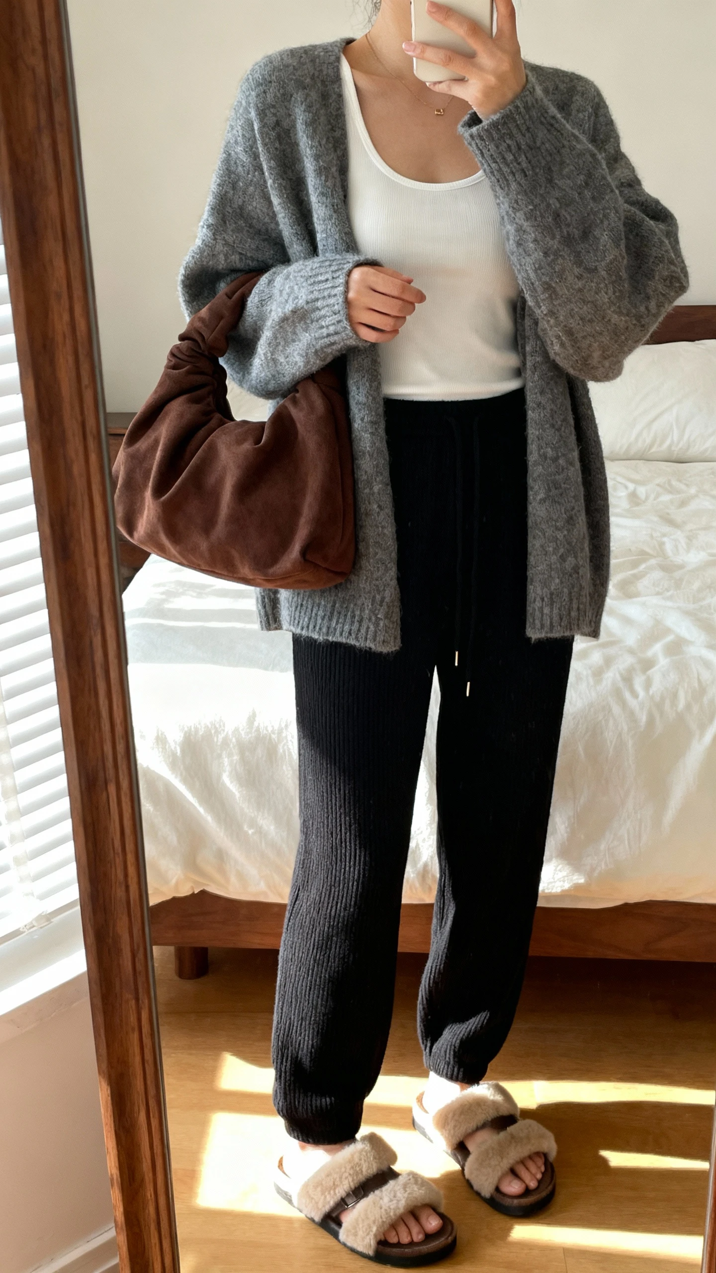 iPhone mirror selfie of a woman wearing a soft slouchy hobo bag in chocolate, oversized heather-gray cashmere cardigan over a fitted white tank, relaxed black knit joggers, and shearling-lined slide sandals; face not visible, cropped at shoulders, cozy bedroom mirror, warm afternoon light, iPhone photo quality.
