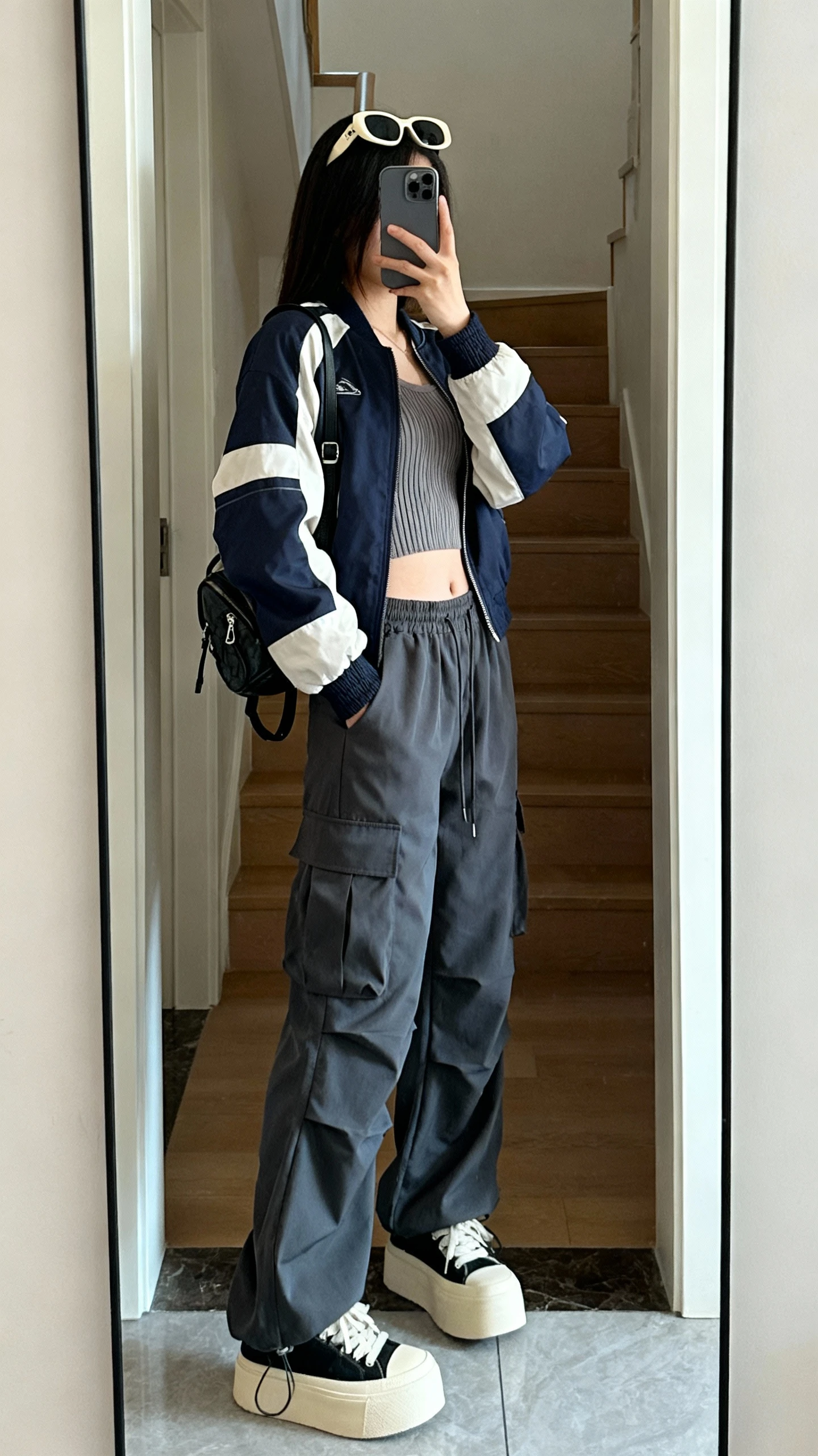 iPhone mirror selfie of a woman wearing a sporty bomber jacket with parachute pants, a ribbed crop top, platform sneakers, chunky sunglasses on head, and a mini backpack, face not visible, side angle, entryway full-length mirror, natural daylight, iPhone photo quality.
