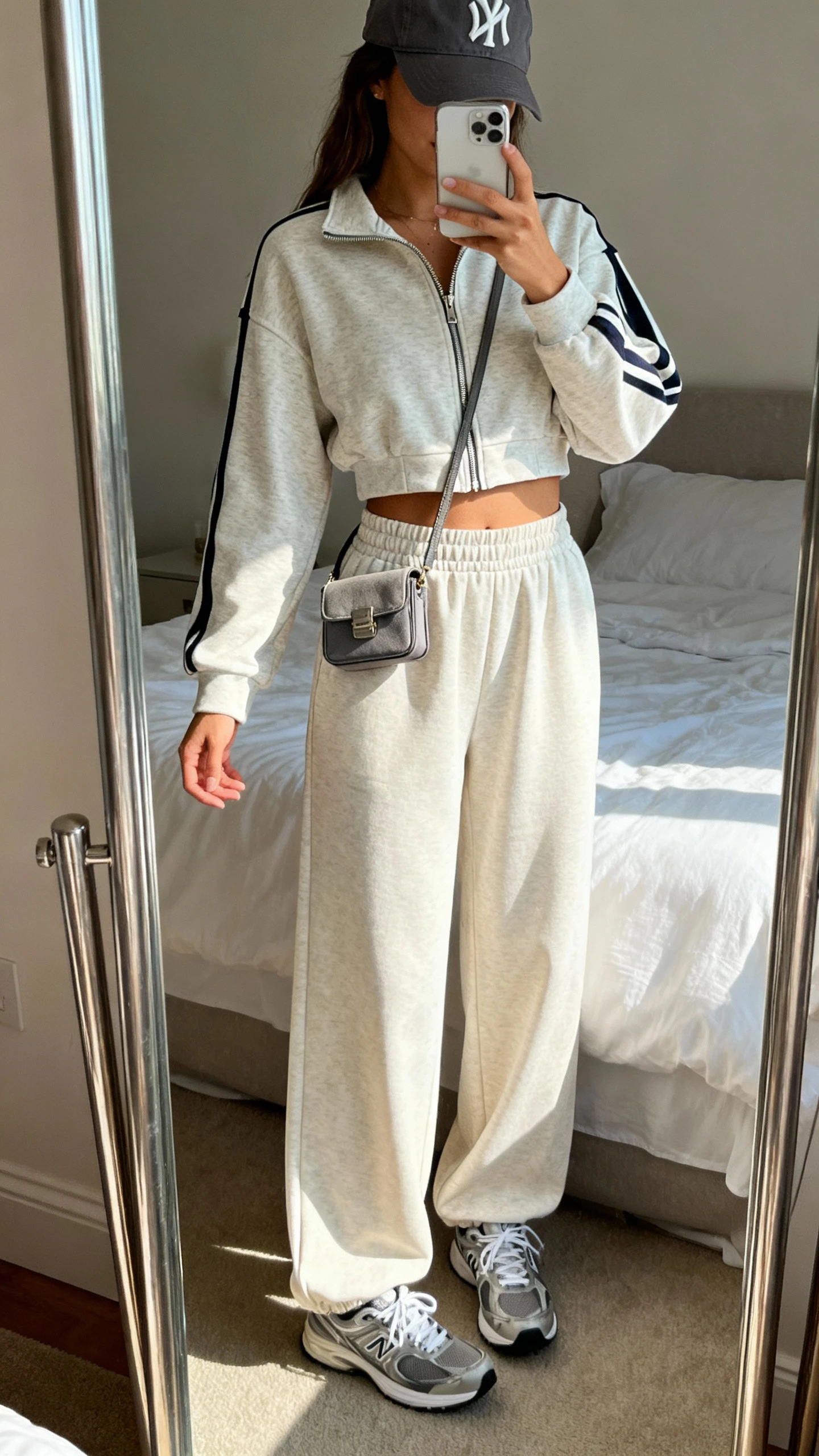 iPhone mirror selfie of a woman wearing a sporty-chic matching set (cropped zip-up track jacket and high-waist wide-leg pants) with sleek trainers, a baseball cap, and a tiny crossbody, face not visible, cropped at shoulders, bedroom mirror, natural daylight, iPhone photo quality.