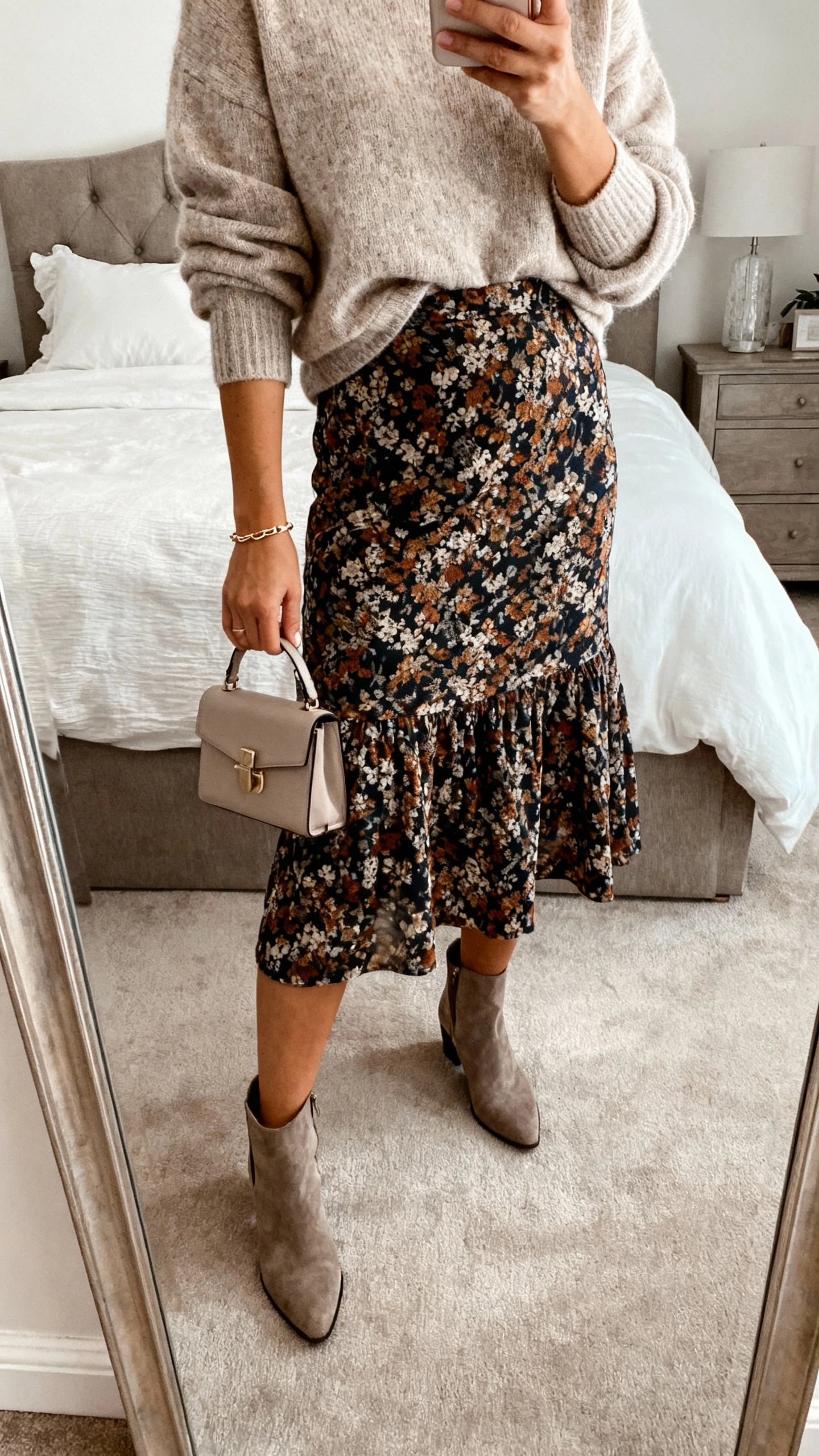 iPhone mirror selfie of a woman wearing a statement skirt with a soft sweater half-tucked, delicate bracelet, small handbag, and ankle boots; face not visible, cropped at neck, bedroom mirror with diffused daylight, iPhone photo quality.