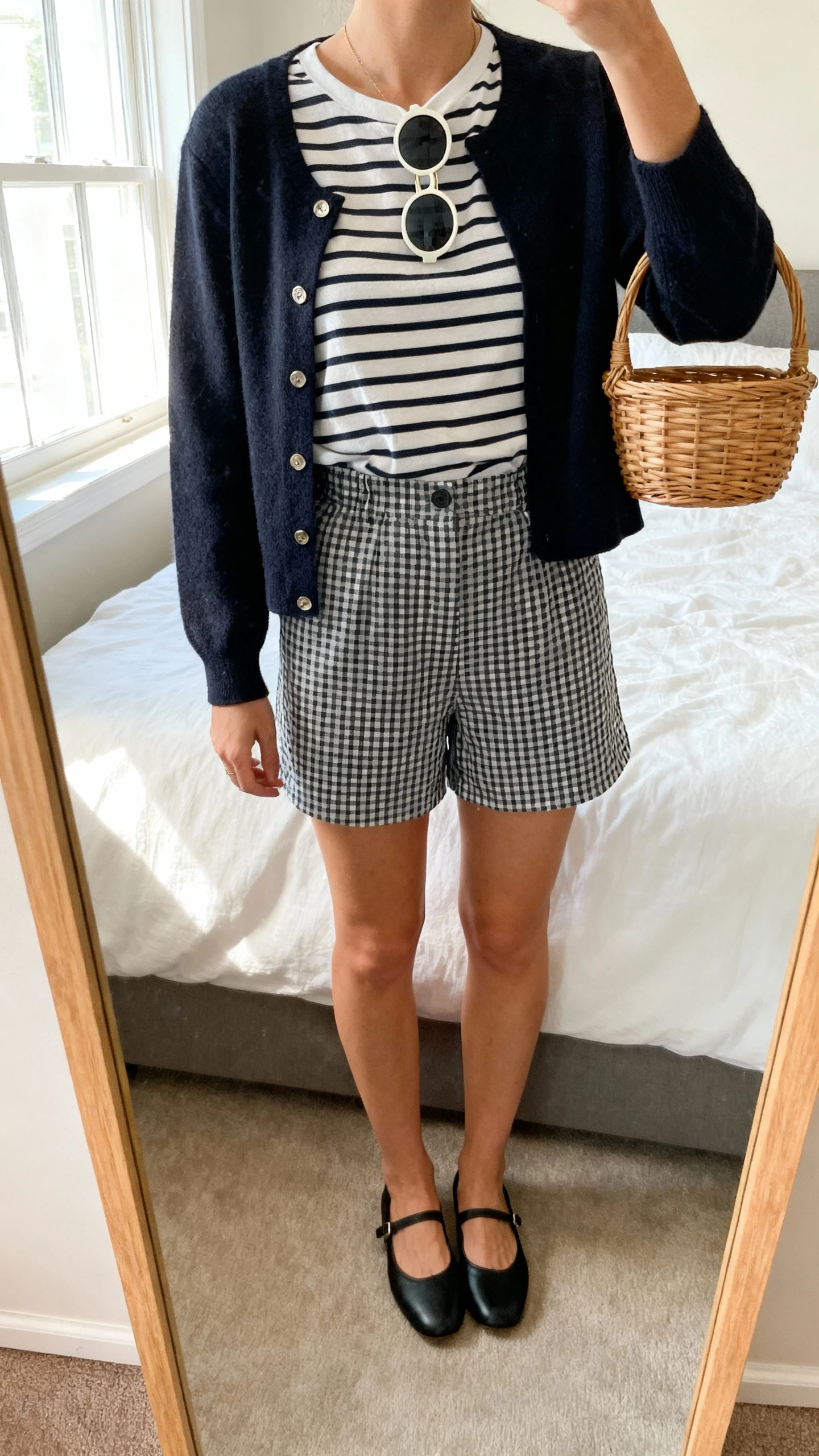 iPhone mirror selfie of a woman wearing a striped Breton tee tucked into high-waisted gingham shorts, black ballet flats, a cropped mod cardigan, round sunglasses hooked on the neckline, and a small wicker basket bag, face not visible, cropped at neck, bedroom mirror with morning window light, iPhone photo quality.