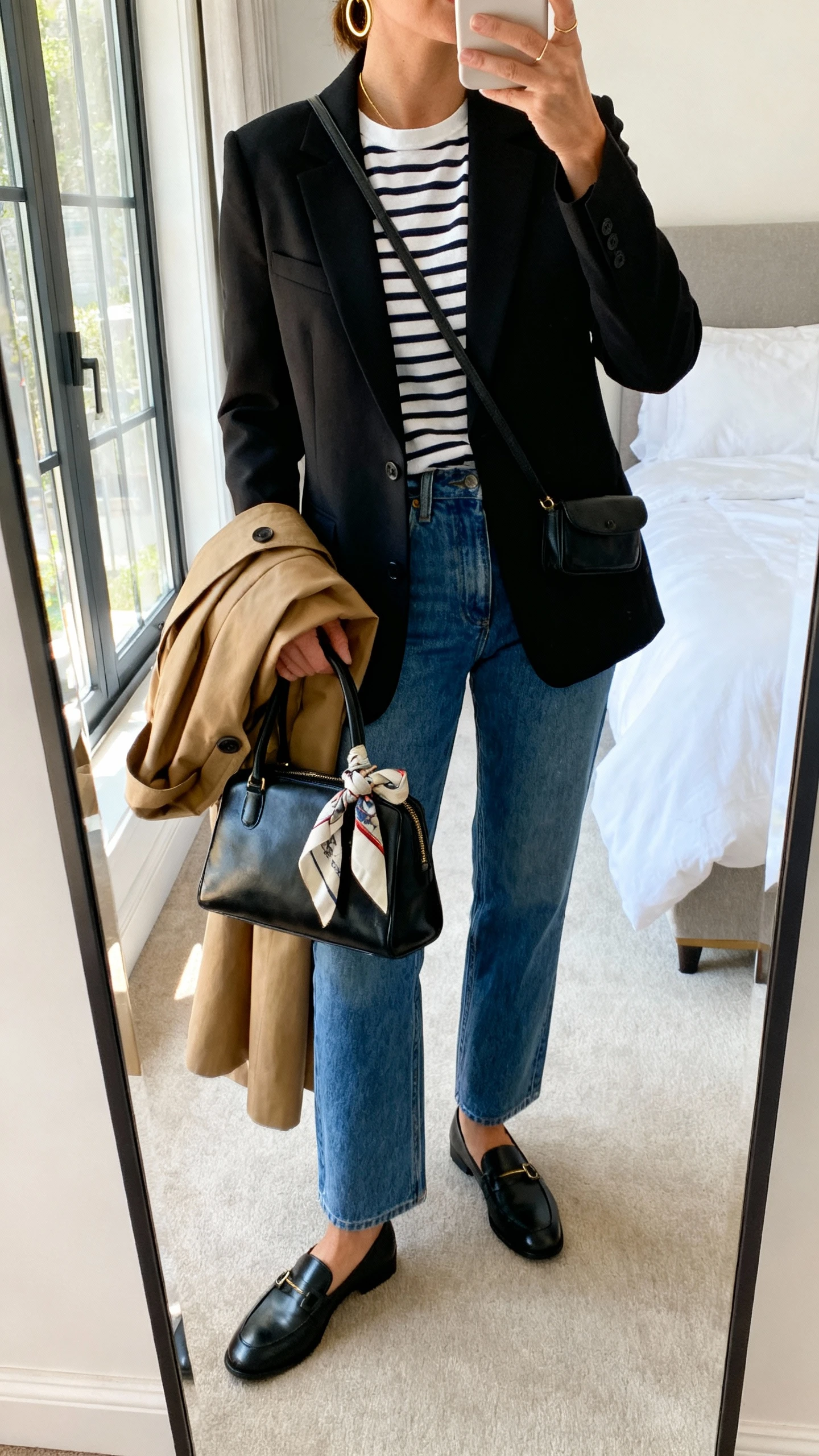 iPhone mirror selfie of a woman wearing a striped Breton tee, tailored black blazer, straight-leg blue jeans, black loafers, tan trench coat draped over arm, black leather crossbody, thin gold hoops, and a silk scarf tied to the bag handle, face not visible, cropped at neck, bedroom mirror, natural window light, iPhone photo quality.