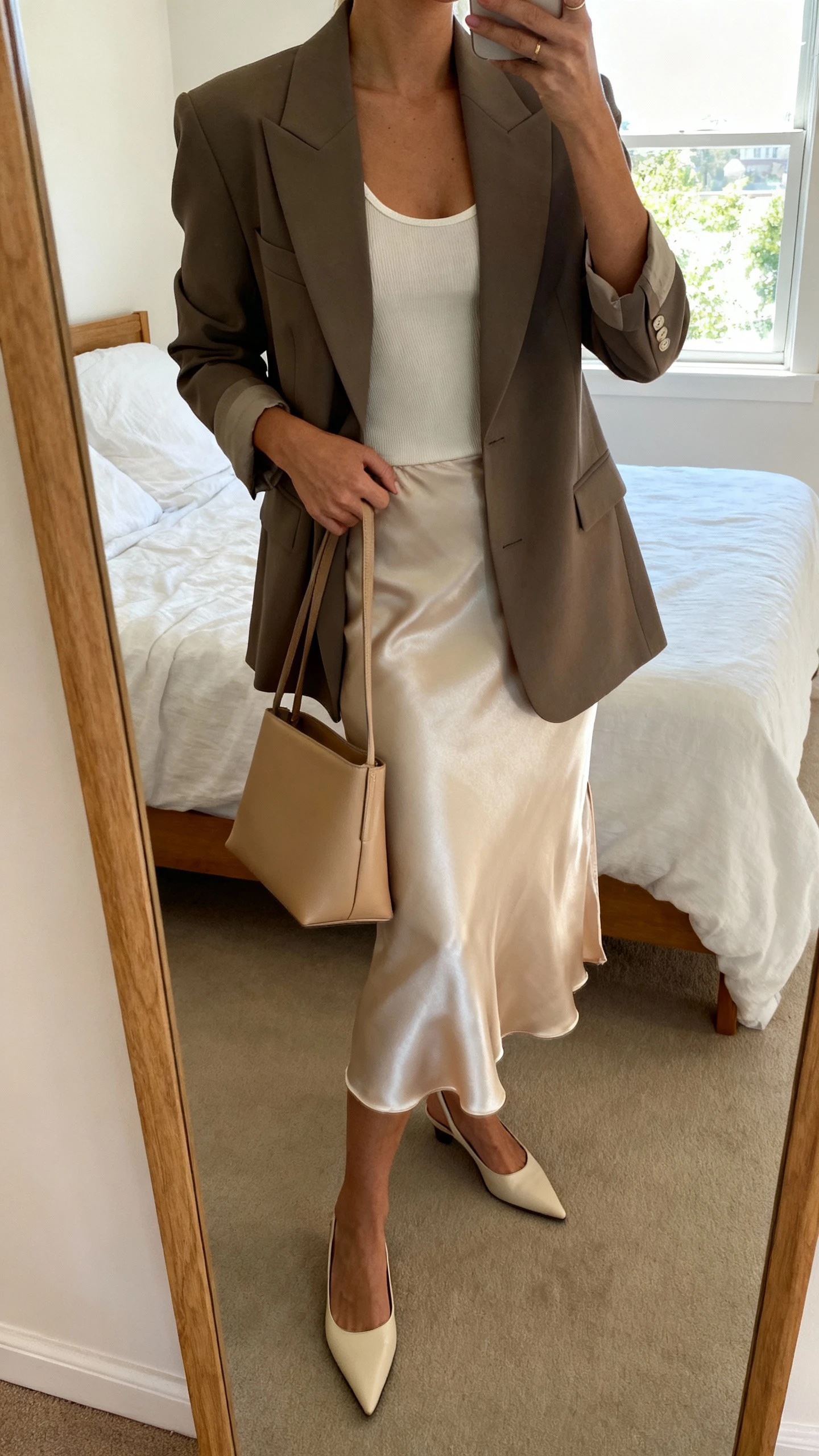 iPhone mirror selfie of a woman wearing a structured power blazer over a silky slip skirt with a fitted tank, pointed-toe heels, and a minimalist leather tote, face not visible, cropped at neck, bedroom mirror, natural window light, iPhone photo quality.