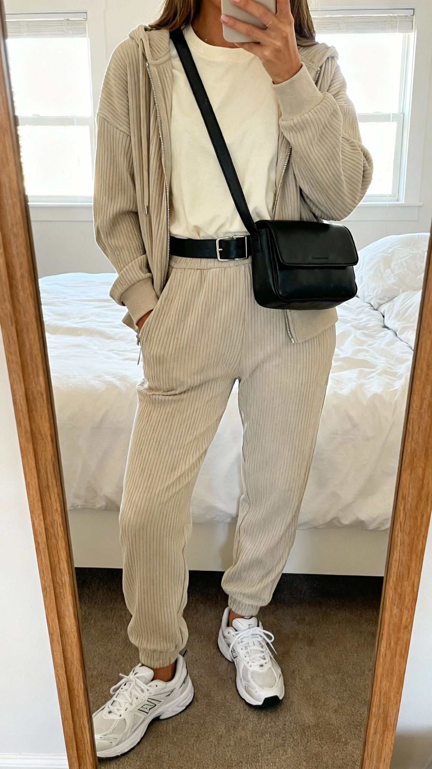 iPhone mirror selfie of a woman wearing a structured black belt bag worn crossbody, a matching ribbed knit zip hoodie and joggers set, a soft cream crewneck tee, and breathable white sneakers, face not visible, cropped at neck, bedroom mirror, natural window light, iPhone photo quality.