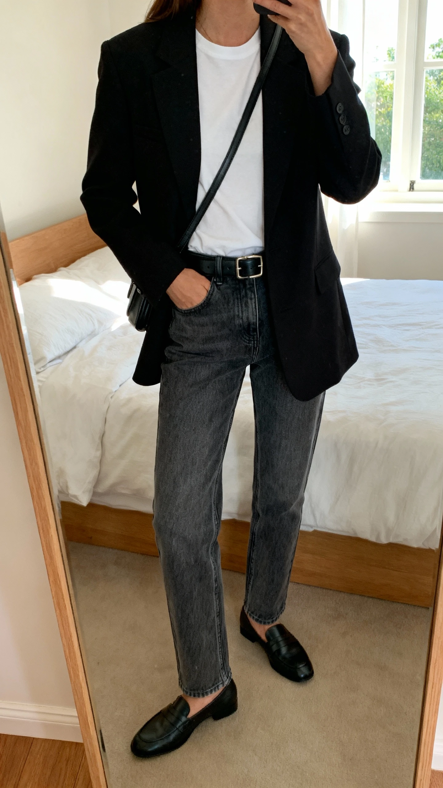 iPhone mirror selfie of a woman wearing a structured black blazer over a white tee, straight-leg mid-wash jeans, black leather belt, loafers, and a simple crossbody bag, face not visible, cropped at neck, bedroom mirror with natural window light, iPhone photo quality.
