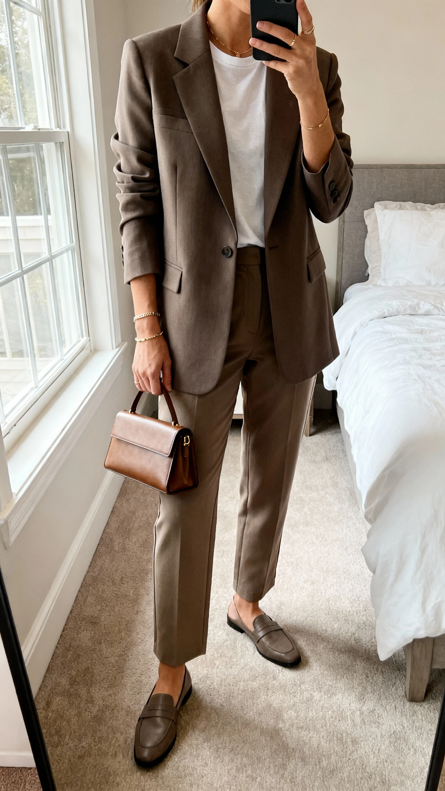 iPhone mirror selfie of a woman wearing a structured power blazer over a simple tee with easy straight-leg trousers and loafers, minimal jewelry and a slim leather tote, face not visible, cropped at neck, bedroom mirror, natural window light, iPhone photo quality.