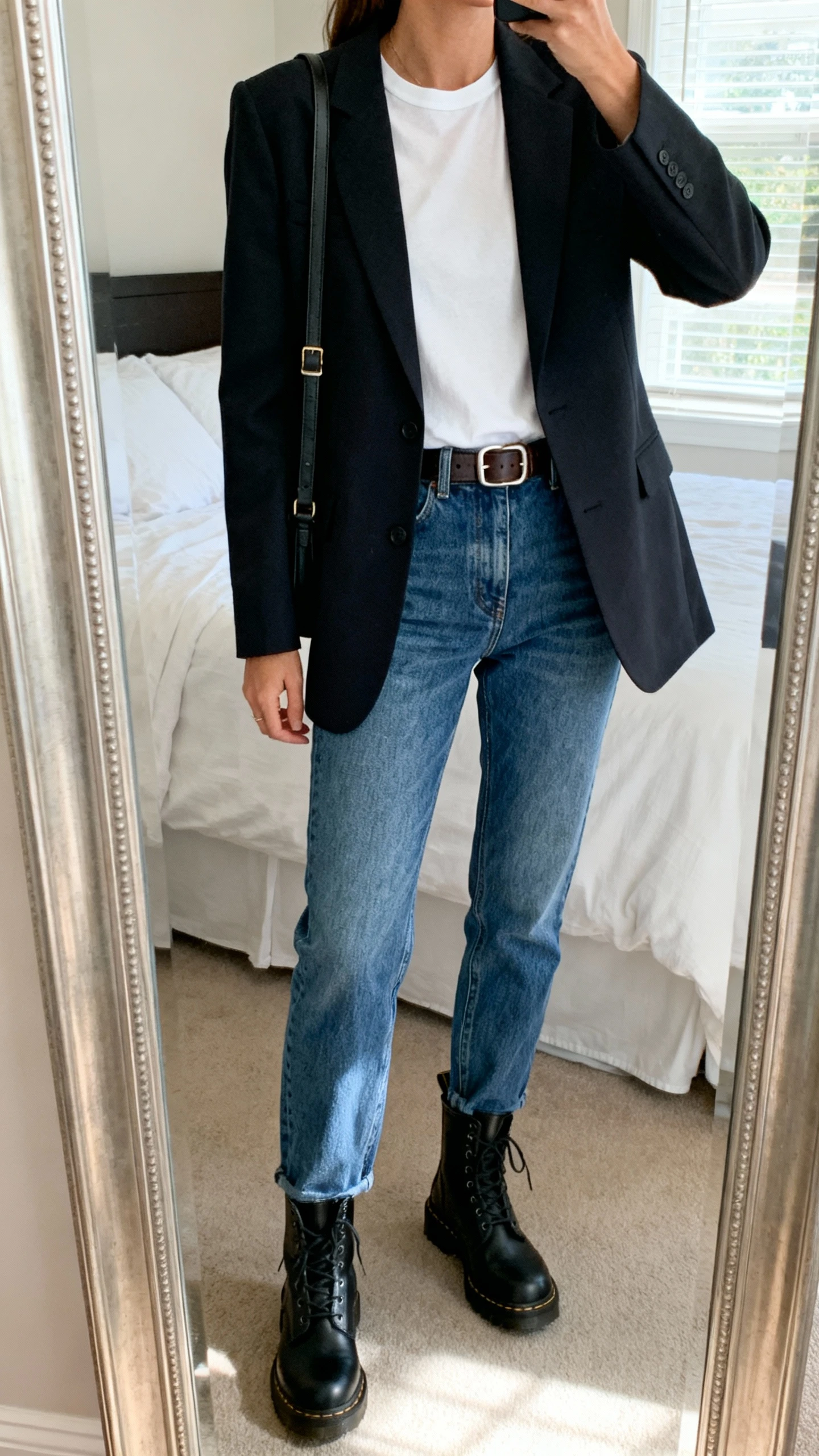 iPhone mirror selfie of a woman wearing a tailored blazer over a classic white tee, straight-leg blue denim, black combat boots, simple leather belt, and a crossbody bag, face not visible, cropped at neck, bedroom mirror, natural window light, iPhone photo quality.