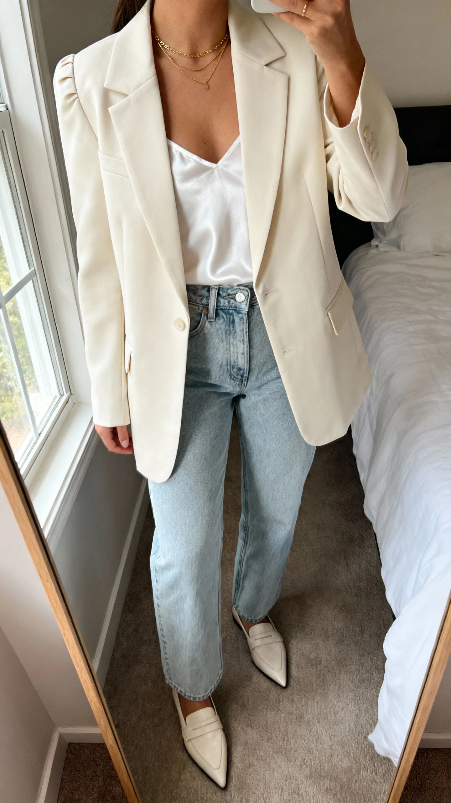 iPhone mirror selfie of a woman wearing a tailored cream blazer with subtle shoulders, high-waisted straight-leg light-wash jeans, a white silky camisole, pointed-toe loafers, and small gold layered necklaces; face not visible, cropped at neck, bedroom mirror with morning window light, iPhone photo quality.