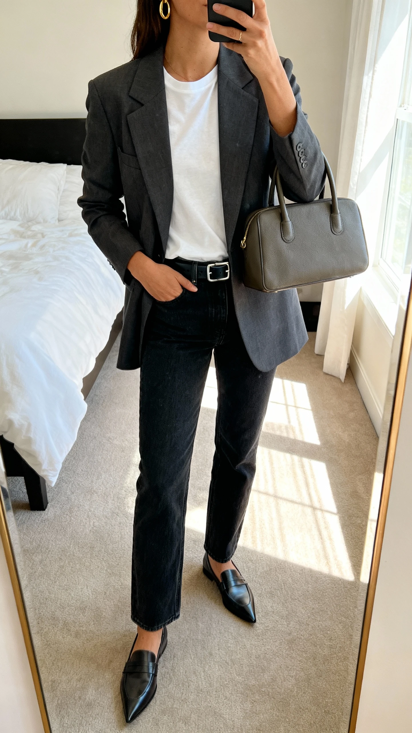 iPhone mirror selfie of a woman wearing a tailored blazer over a crisp white tee, straight-leg dark jeans, sleek belt, pointed-toe loafers, minimal gold hoops, structured top-handle bag, face not visible, cropped at neck, bedroom mirror, morning window light, iPhone photo quality.
