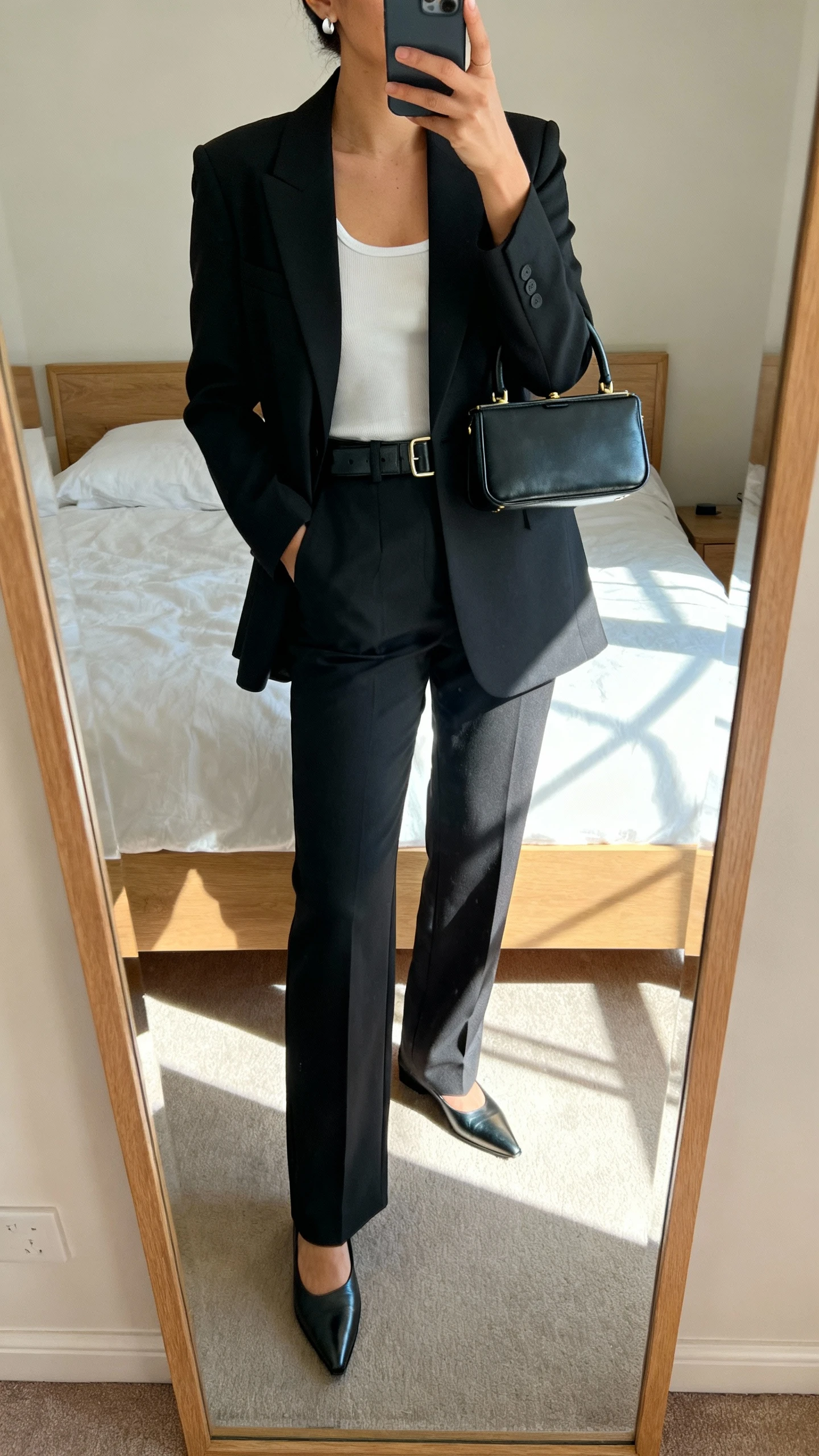 iPhone mirror selfie of a woman wearing a tailored monochrome set: sharp blazer, matching straight-leg trousers, minimalist tank, sleek belt, pointed loafers, delicate stud earrings, structured mini top-handle bag; face not visible, cropped at neck, bedroom mirror with morning window light, iPhone photo quality.