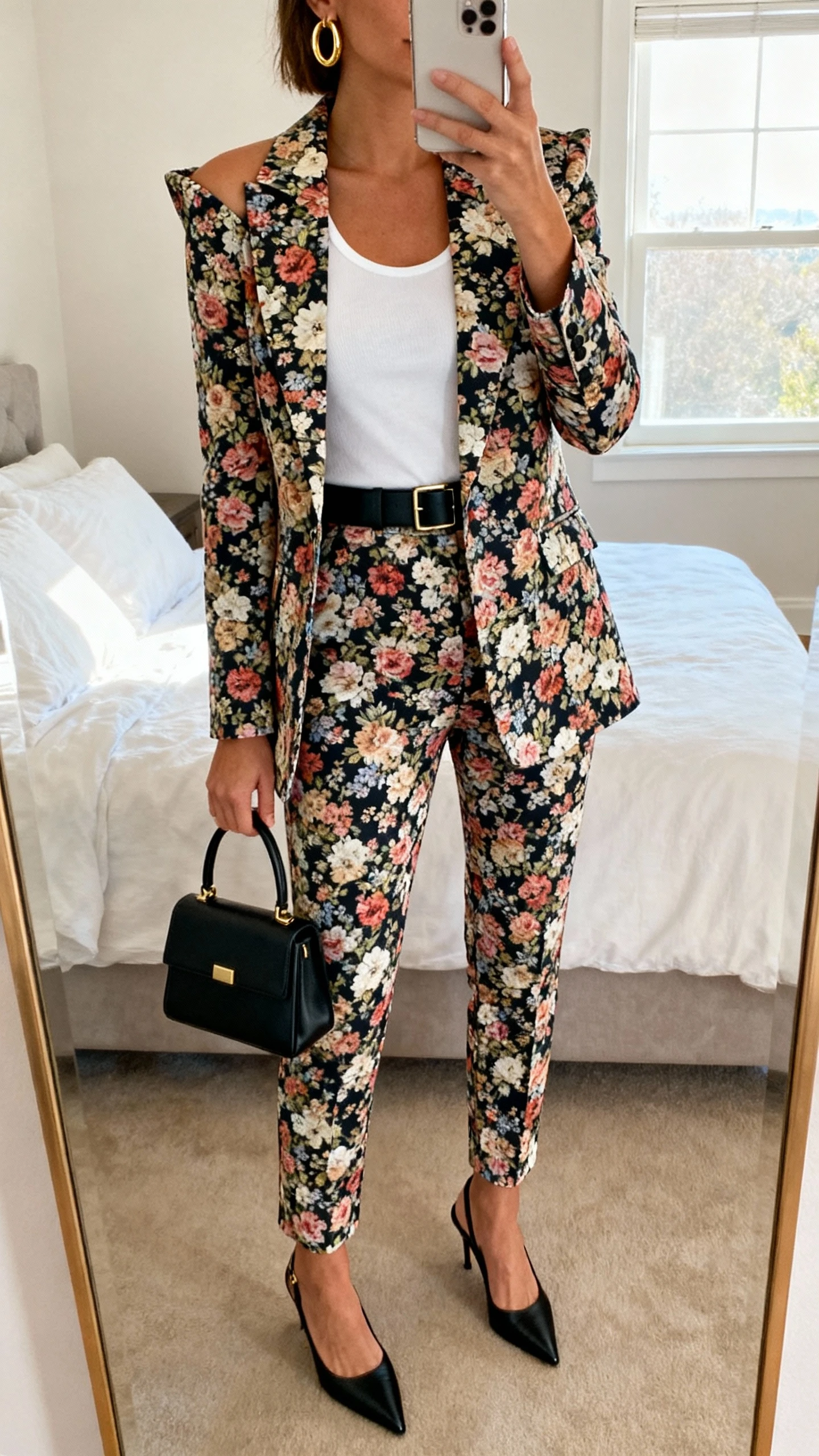 iPhone mirror selfie of a woman wearing a tailored floral power suit (sharp-shoulder blazer with matching tapered trousers) over a crisp white tank, pointed black slingback heels, slim black belt, structured mini top-handle bag, and gold hoop earrings; face not visible, cropped at neck, bedroom mirror, natural morning window light, iPhone photo quality.