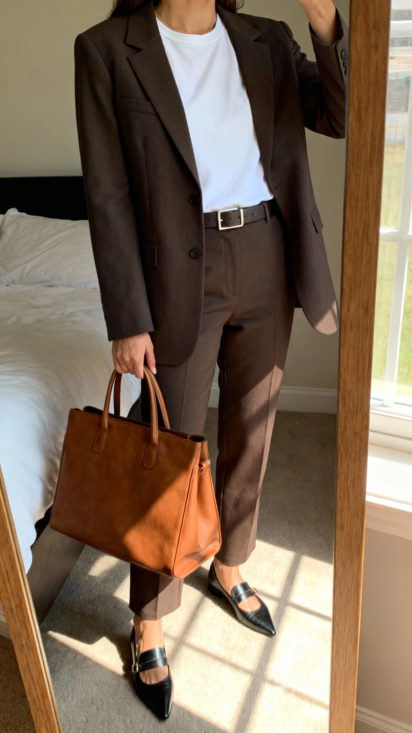 iPhone mirror selfie of a woman wearing a tailored blazer over a crisp white perfect tee, straight-leg trousers, pointed loafers, a structured leather tote, and a slim belt; face not visible, cropped at neck, bedroom mirror with morning window light, iPhone photo quality.