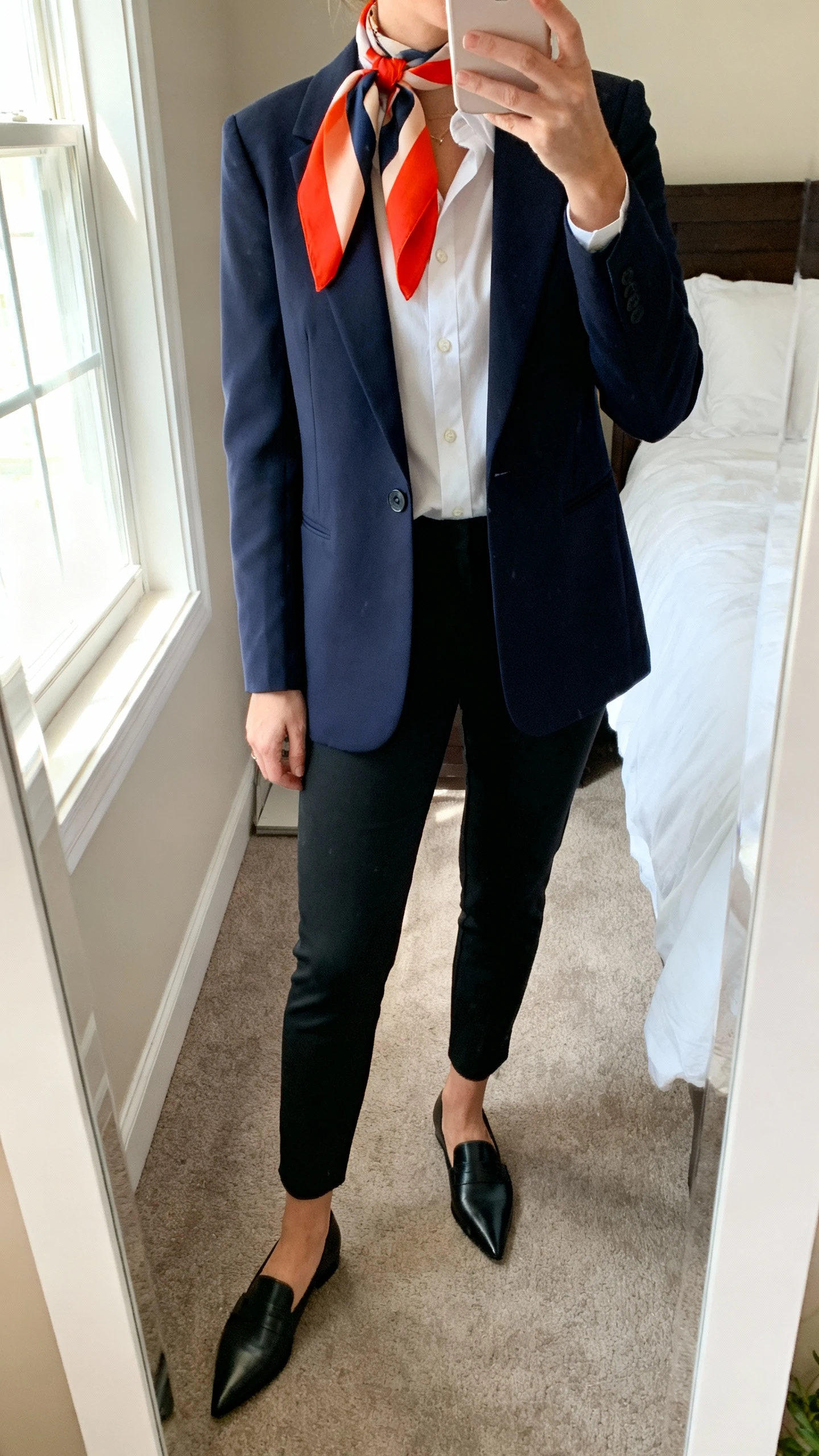 iPhone mirror selfie of a woman wearing a tailored navy blazer over a crisp white button-down, slim black trousers, pointed black loafers, and a bright color-block silk scarf tied at the neck, face not visible, cropped at shoulders, bedroom mirror, morning window light, iPhone photo quality.
