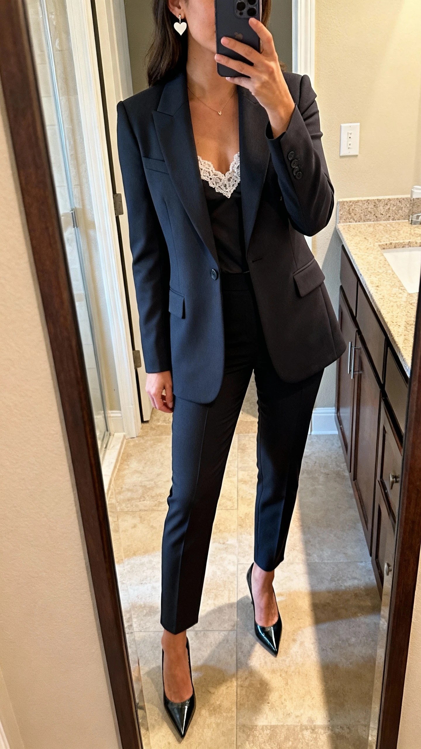 iPhone mirror selfie of a woman wearing a tailored power suit with a fitted blazer and tapered trousers, lace camisole peeking underneath, dainty heart earrings and pointed-toe pumps, face not visible, bathroom full-length mirror, warm indoor lighting, iPhone photo quality